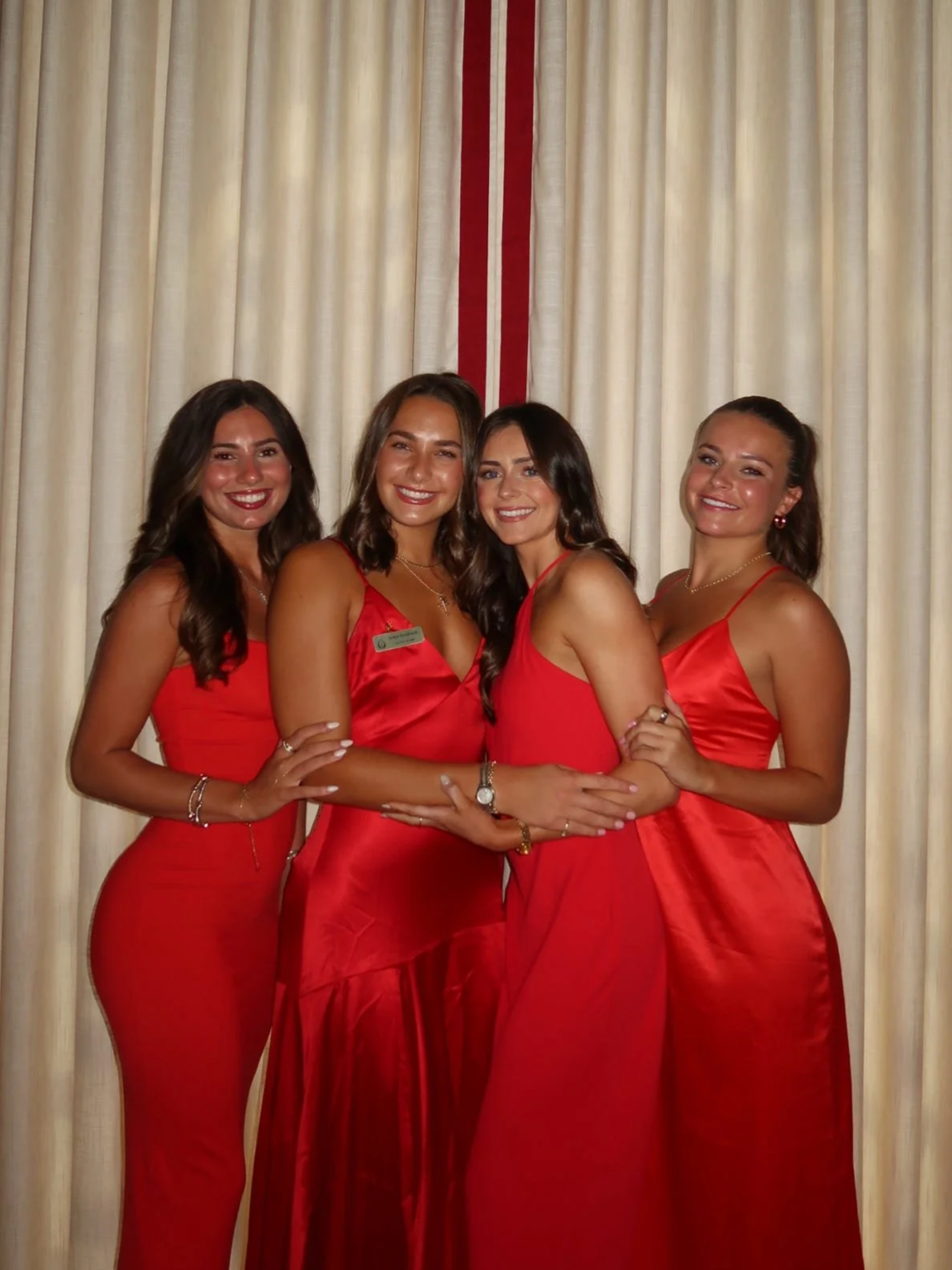 Four women in red dresses standing in front of cream-colored curtains with red vertical ribbon accents, smiling for a photo.