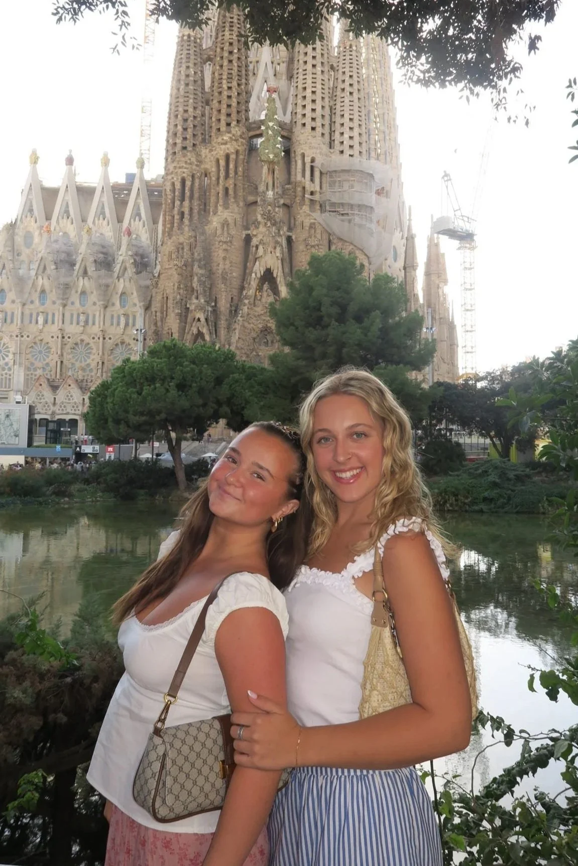 Two young women smiling and posing together in front of a pond with greenery and trees, with the Sagrada Familia basilica under construction in the background in Barcelona, Spain.