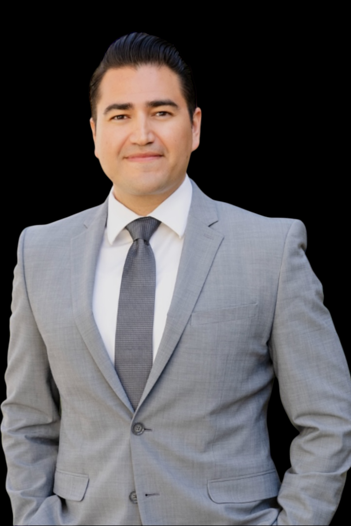 Man in a gray suit with a blue tie, smiling outdoors with greenery in the background.