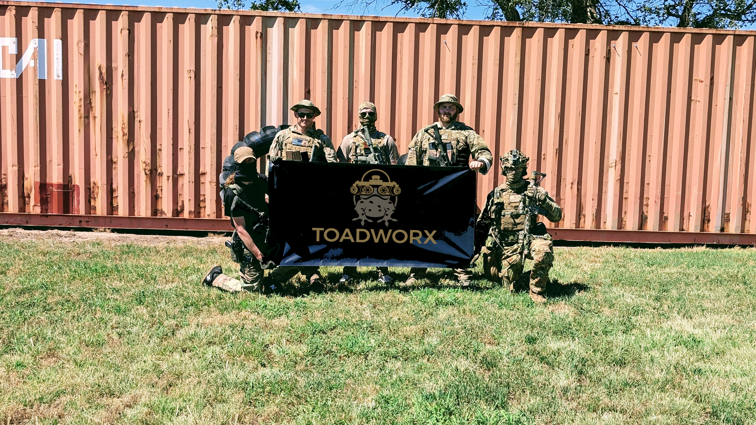 Group of six people in tactical gear holding a black flag with the logo and text 'TOADWORX' in front of a large red shipping container on a grassy field.