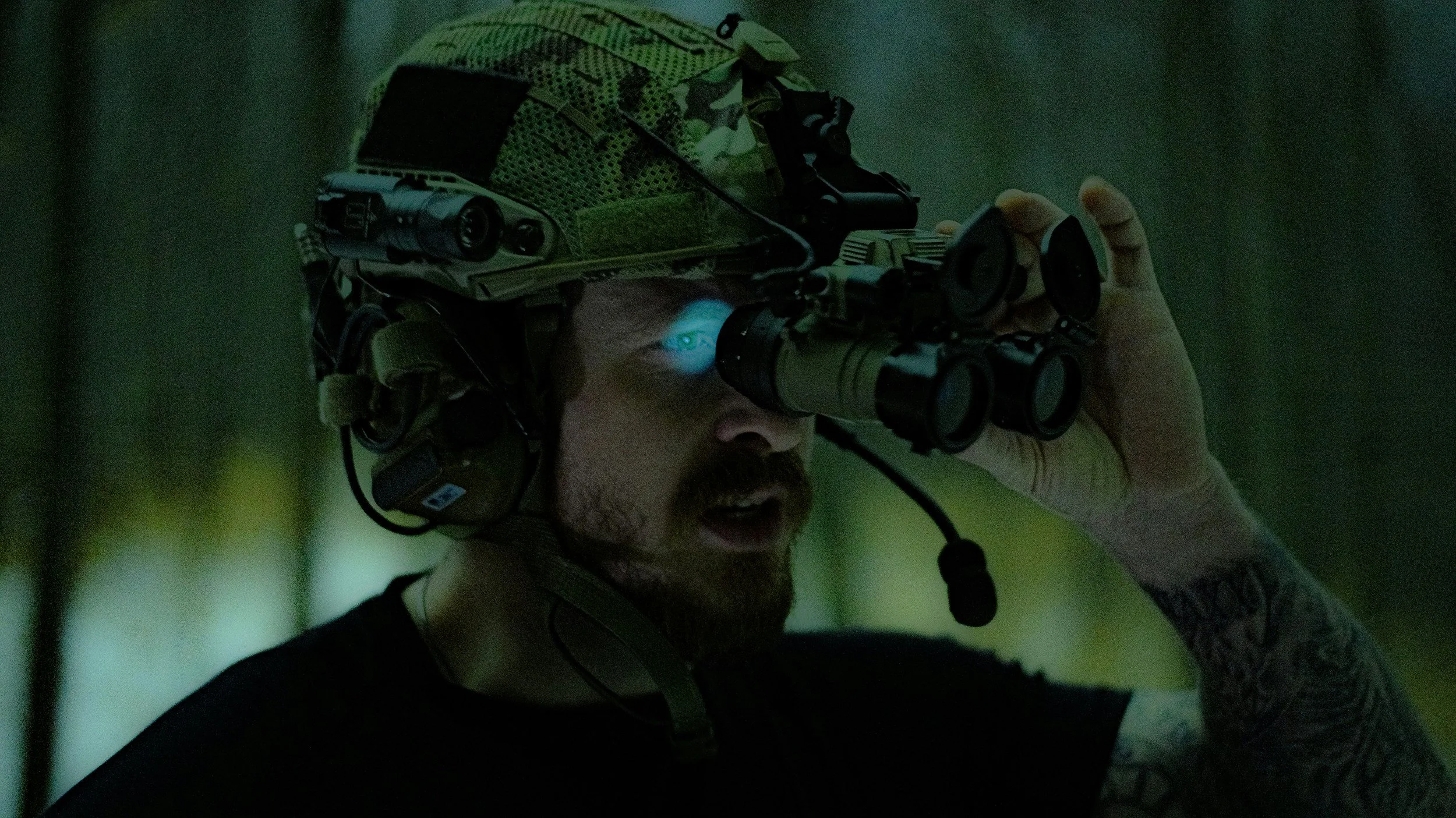 A soldier with a camo helmet and tactical headset looking through binoculars in a dark environment
