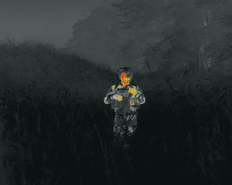 Person walking through tall grass at night, seen with thermal imaging that highlights the person's face and parts of their body in warm colors.