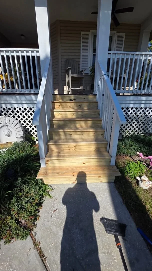 Wooden stairs leading up to a porch with white railings and a chair, surrounded by garden plants, with the shadow of a person and a broom visible on the ground.