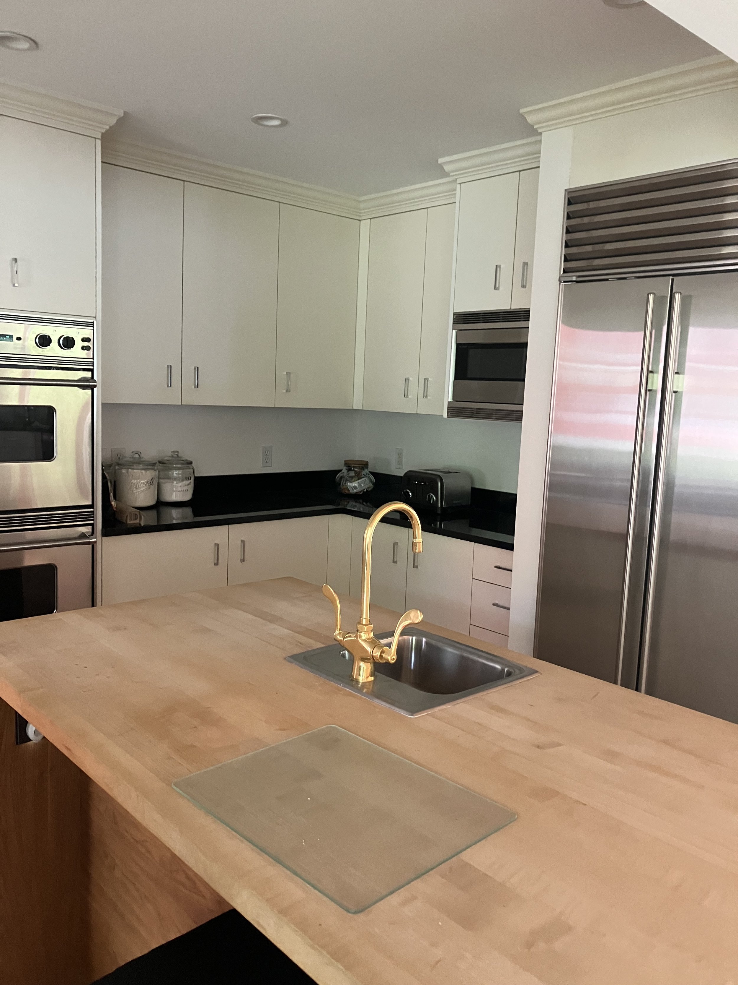 Modern kitchen with white cabinets, stainless steel appliances, a central wooden island with a brass faucet, black countertops, and a large double-door refrigerator.
