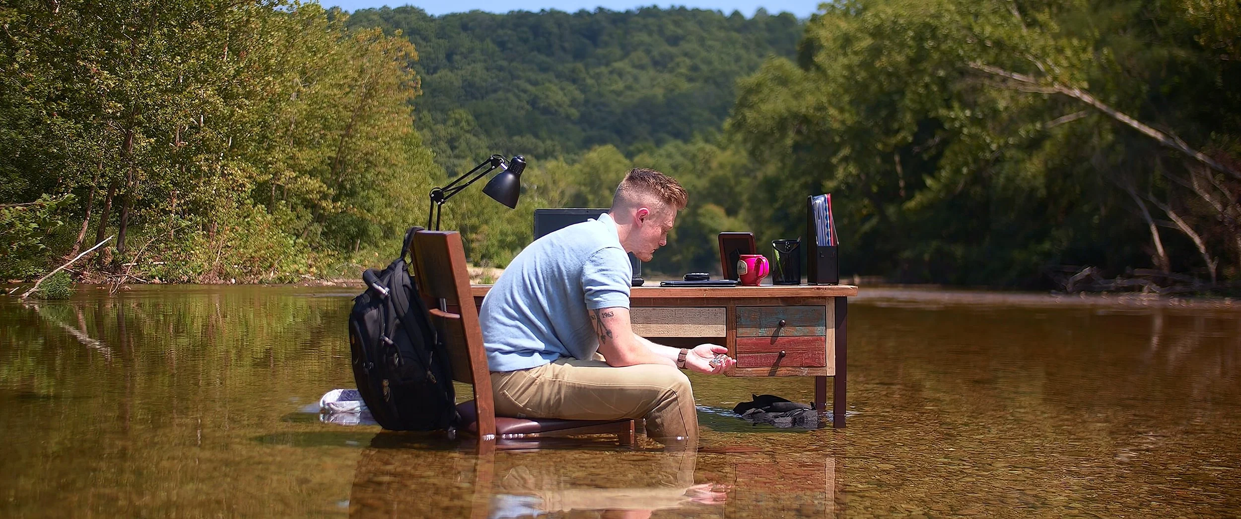 A man sitting at a wooden desk in a river, working on a laptop with books, a mug, and a lamp on the desk, surrounded by green trees and mountains.