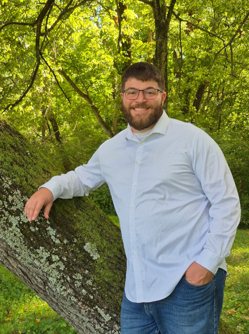 A man with glasses and a beard smiling, leaning on a moss-covered log in a lush green forest.