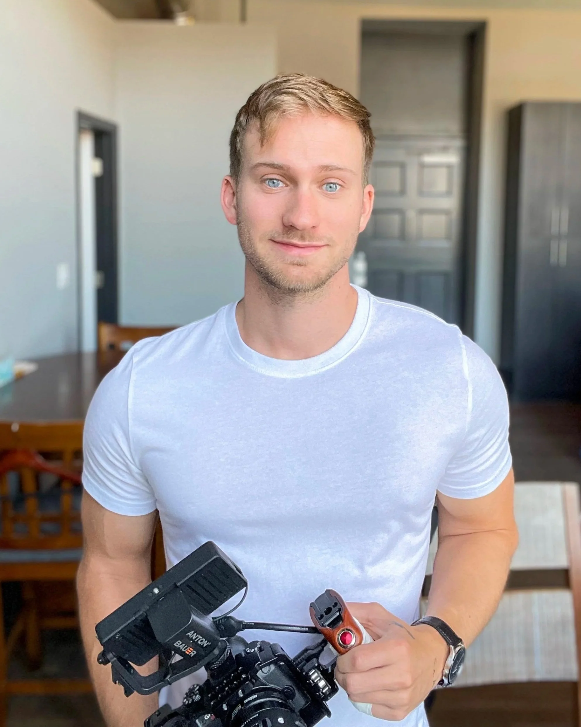 A young man with blonde hair and blue eyes wearing a white t-shirt, holding a professional video camera in a well-lit room.