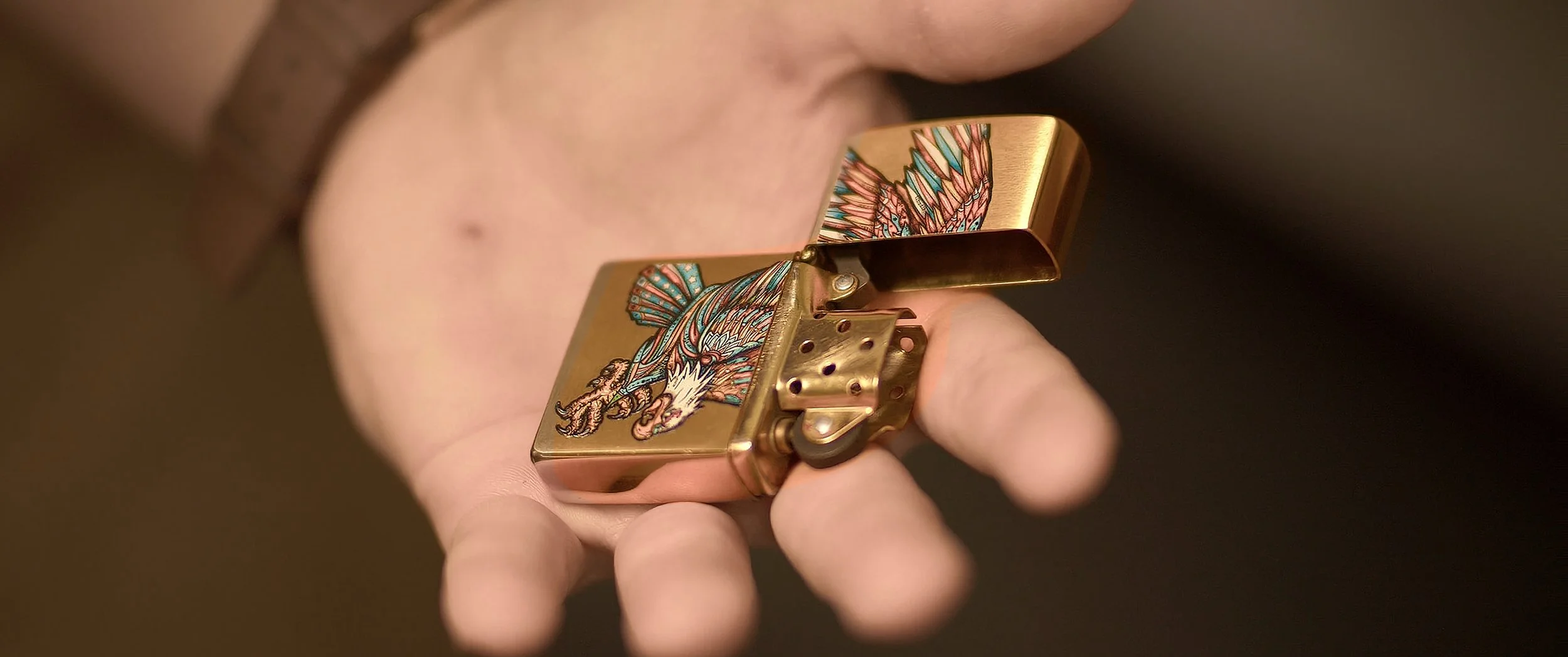 Close-up of a hand holding a gold-colored lighter with an intricate eagle design on the lid.