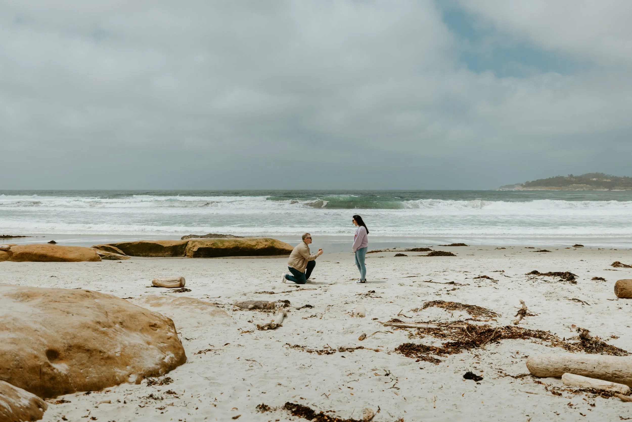 A man proposing to a woman on a Carmel California beach with rocks and ocean in the background.