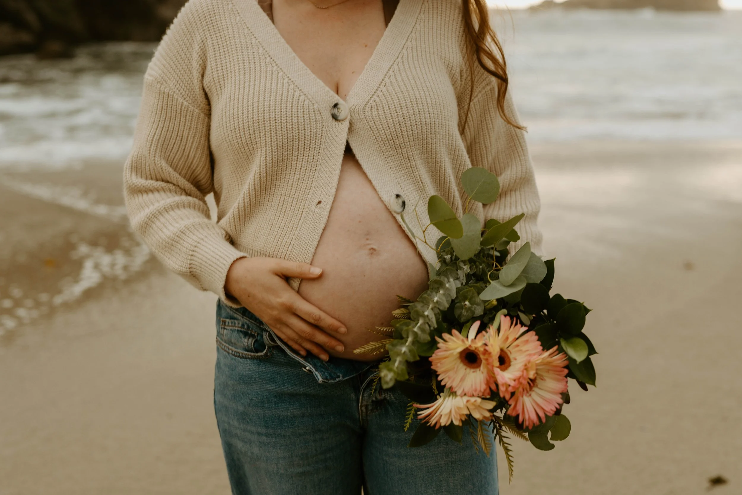 A pregnant woman holding a bouquet of flowers at the beach, wearing a beige cardigan and jeans at Pfeiffer Beach in Big Sur.