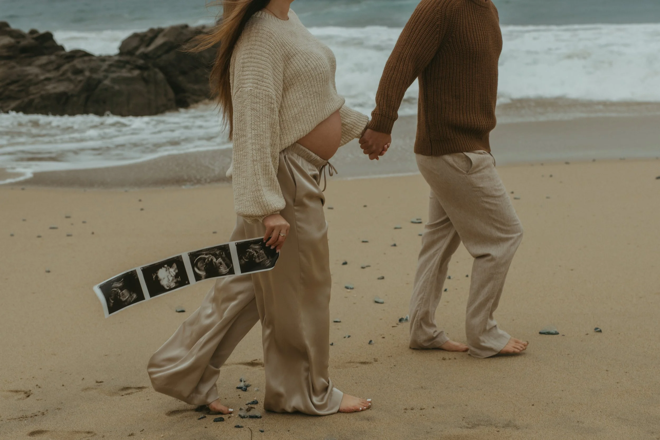 man and pregnant woman walking on Garrapata Beach in Big Sur holding sonogram for maternity photos
