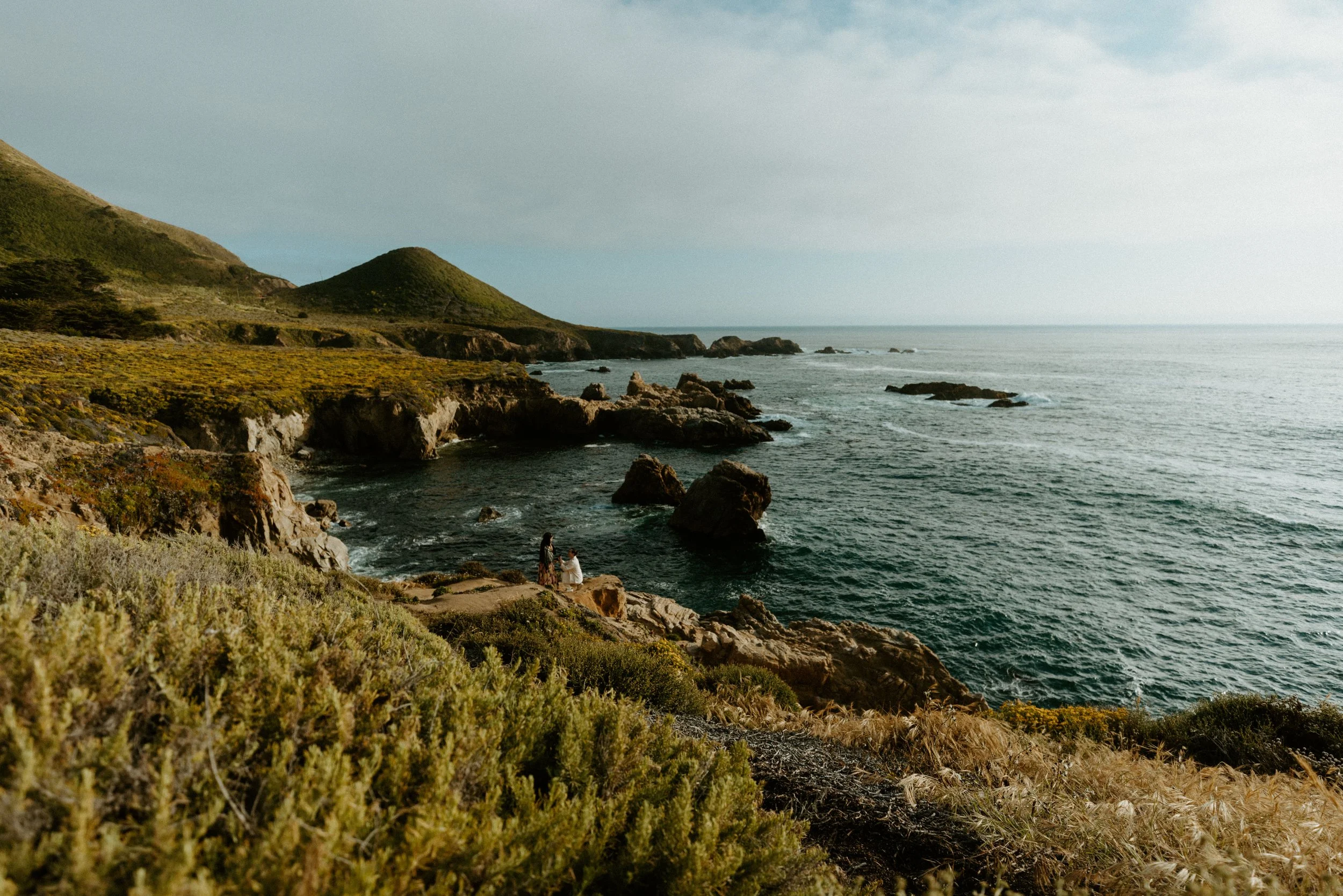 man proposing on one knee with wide angle shot of big sur in the background