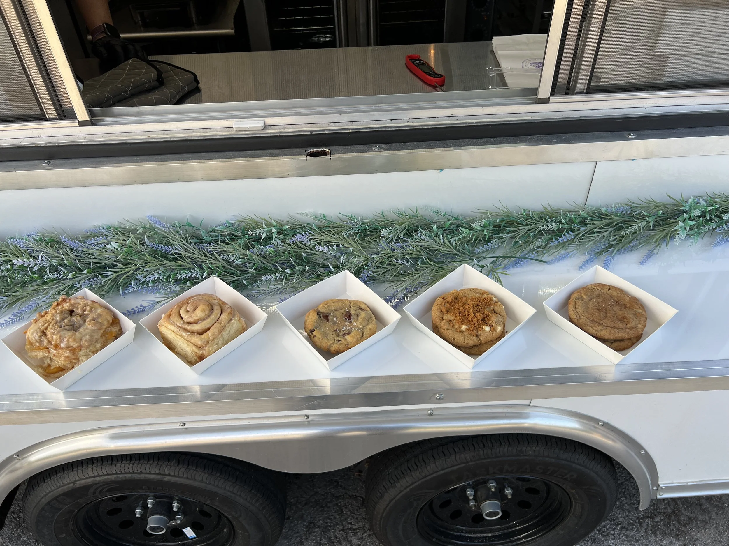 A row of five bakery items in white boxes on a silver cart with purple flowers decoration underneath. The bakery items include a crumble-topped pastry, a cinnamon roll, a chocolate chip cookie, a fudge cookie, and a round cookie or biscuit. The cart is parked outside a building, with a window, some papers, a red tool, and a back entrance visible in the background.