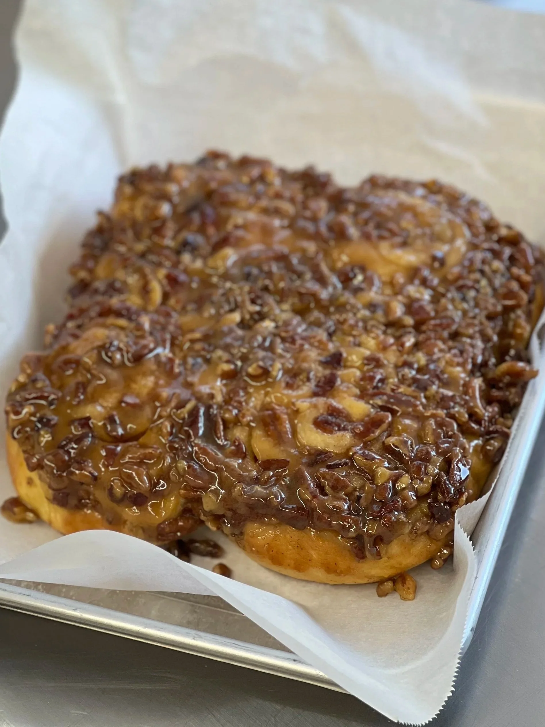 A close-up of a cinnamon roll topped with caramel and pecans in a paper-lined tray.