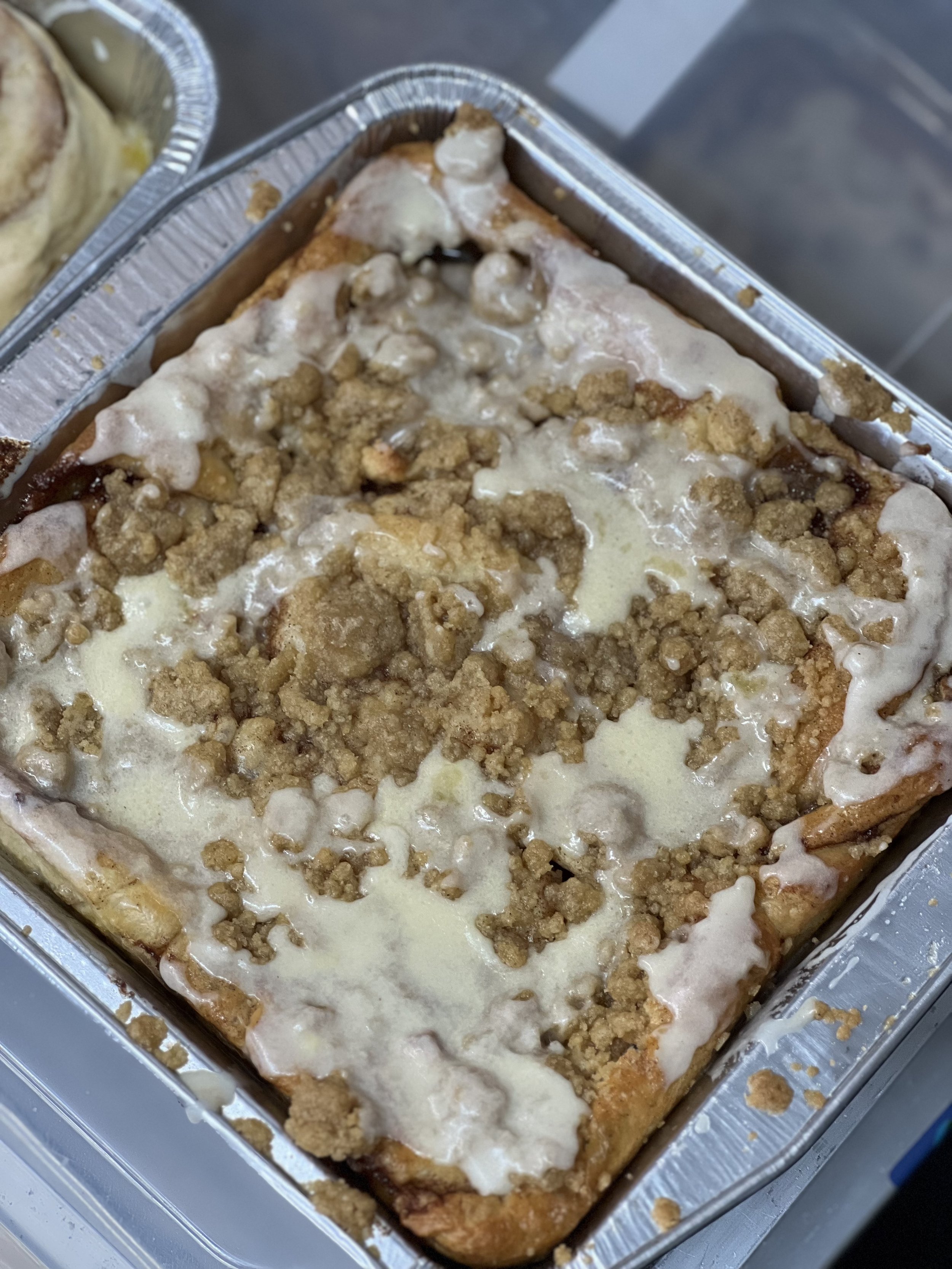 A rectangular dish of baked dessert with a crumbly topping, melted cheese, and white icing drizzled on top.