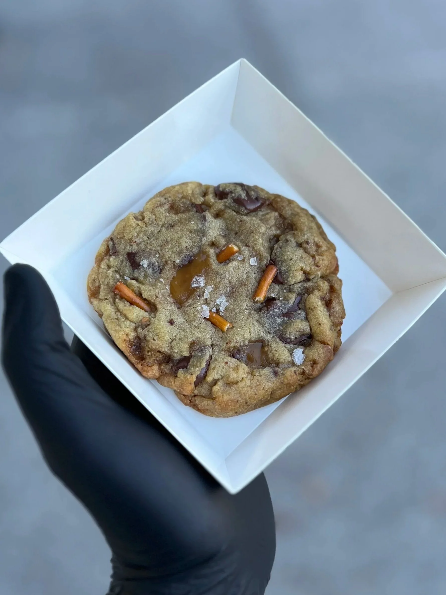 A chocolate chip cookie with salt and pretzel pieces in a white paper container, held by a hand wearing a black glove.