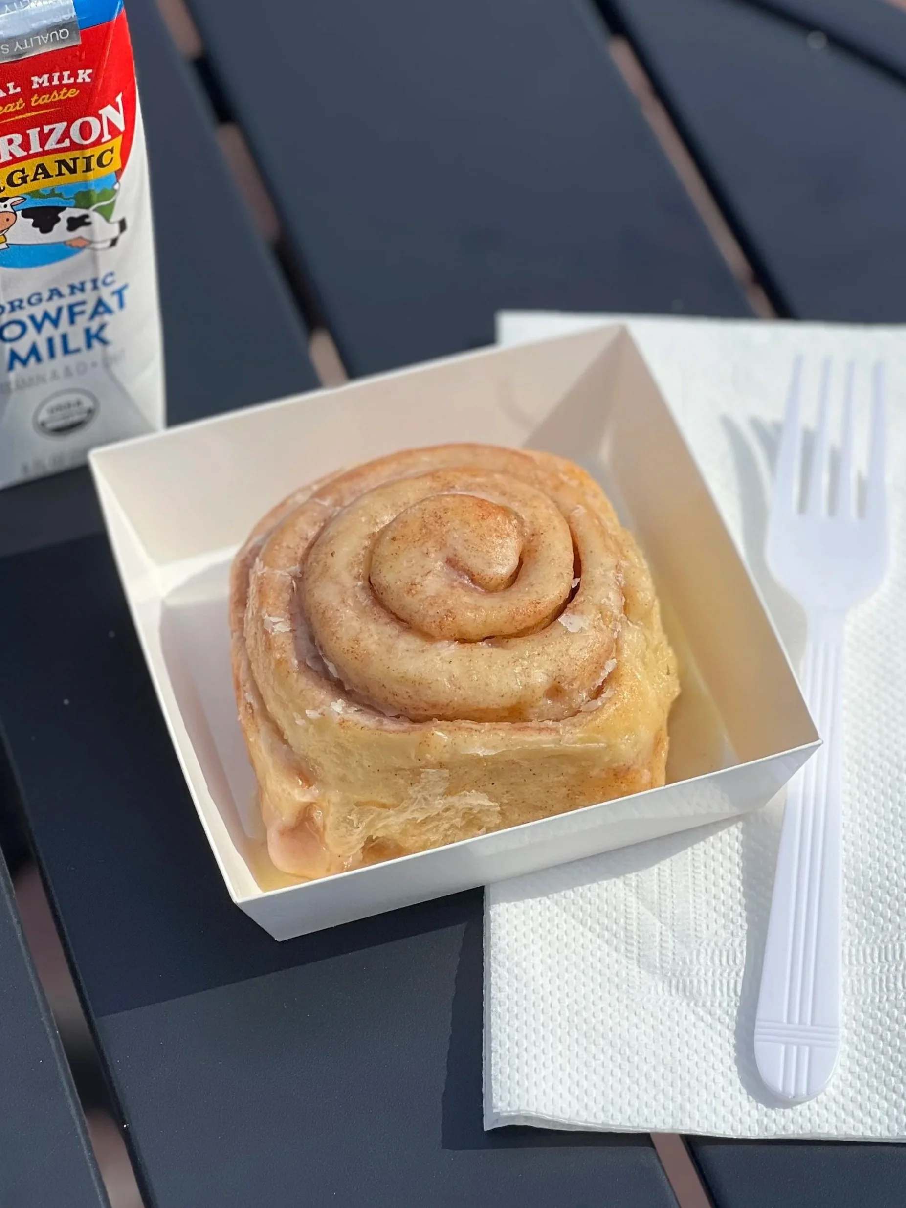 Cinnamon roll in a white paper tray with a fork on a napkin, a carton of organic cow's milk in the background, on an outdoor black table.