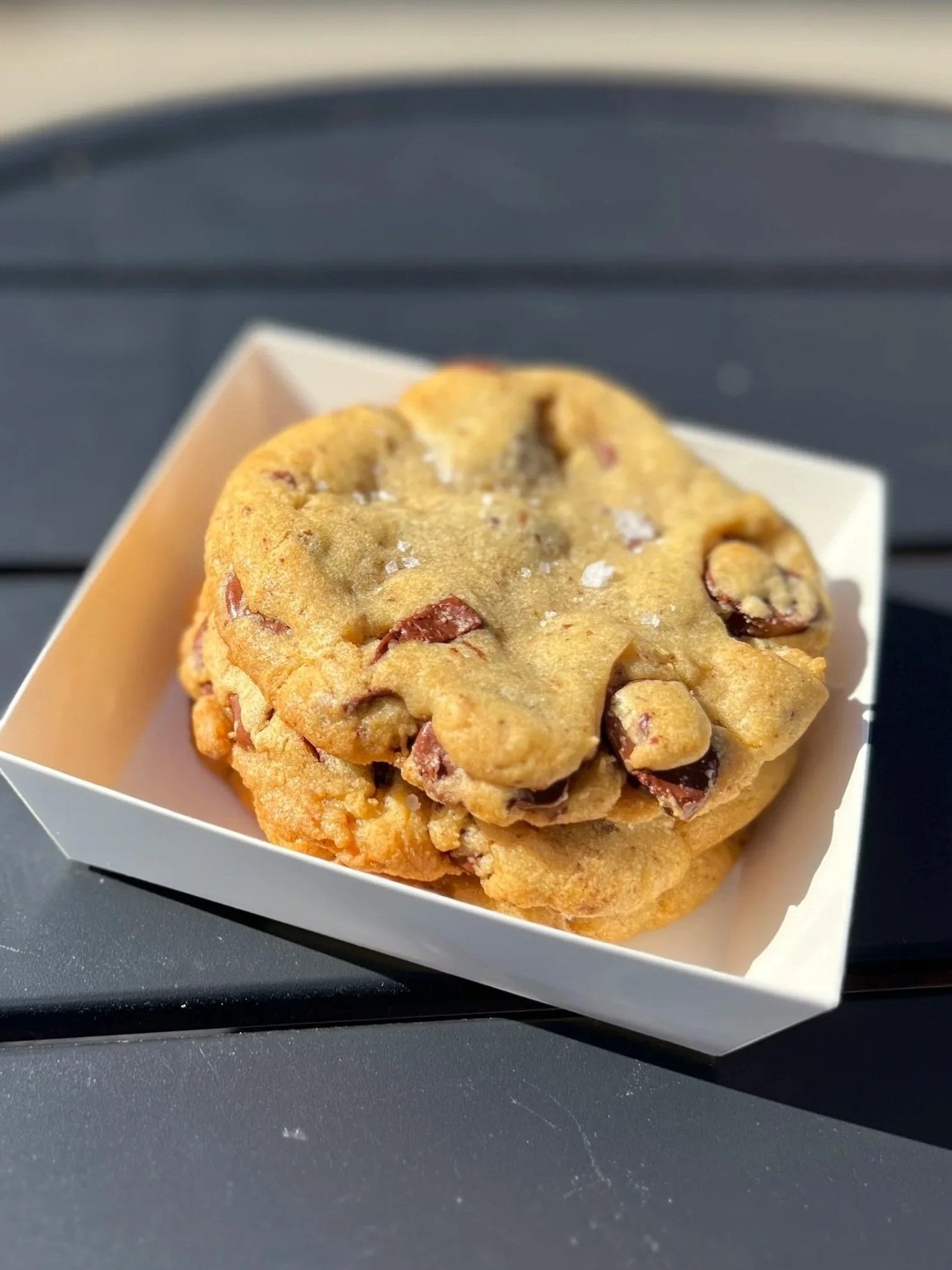 Close-up of a chocolate chip cookie in a white paper tray on a black surface.