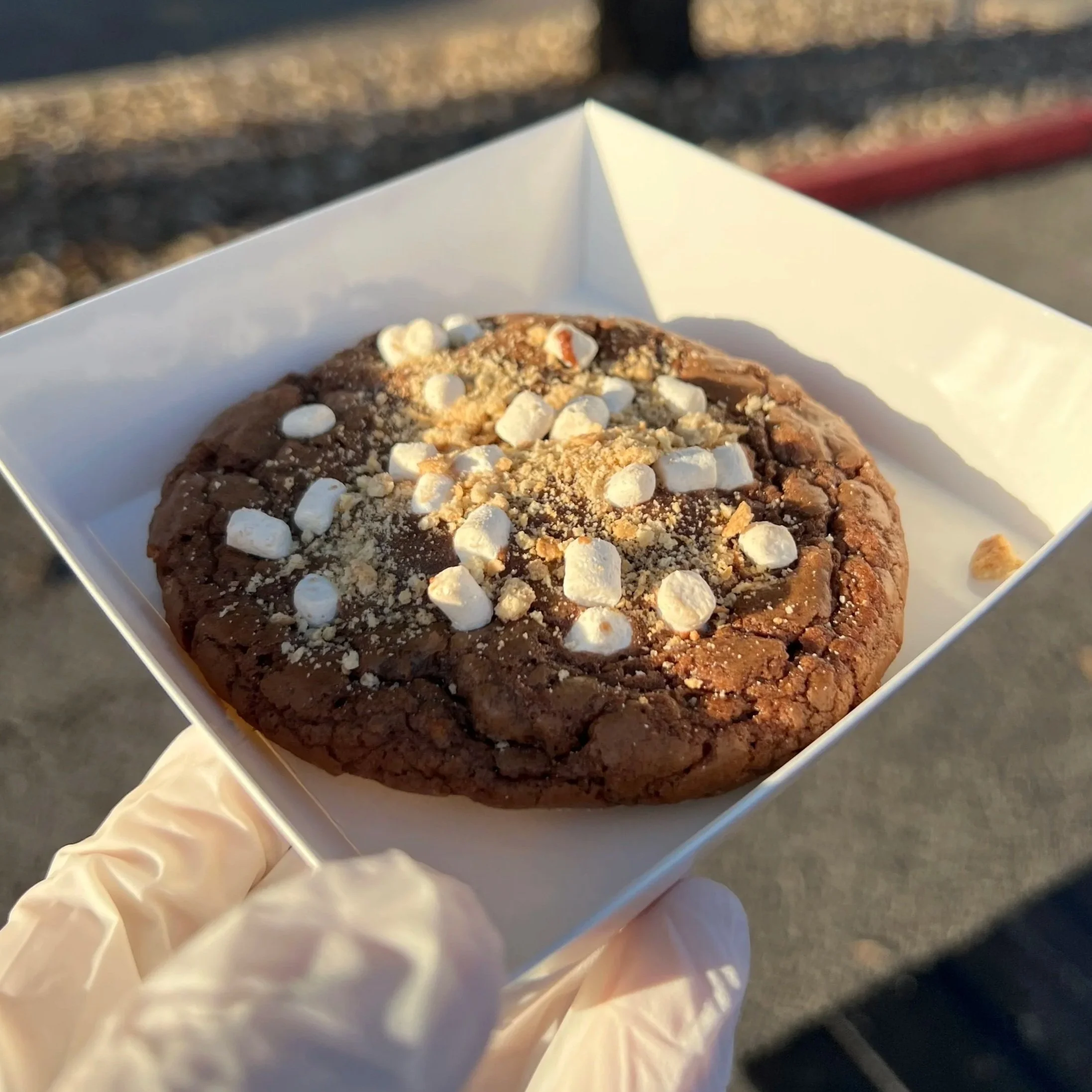 A large chocolate cookie with mini marshmallows and crumbled topping in a white paper tray, held by a person wearing a disposable glove, outdoors near train tracks.