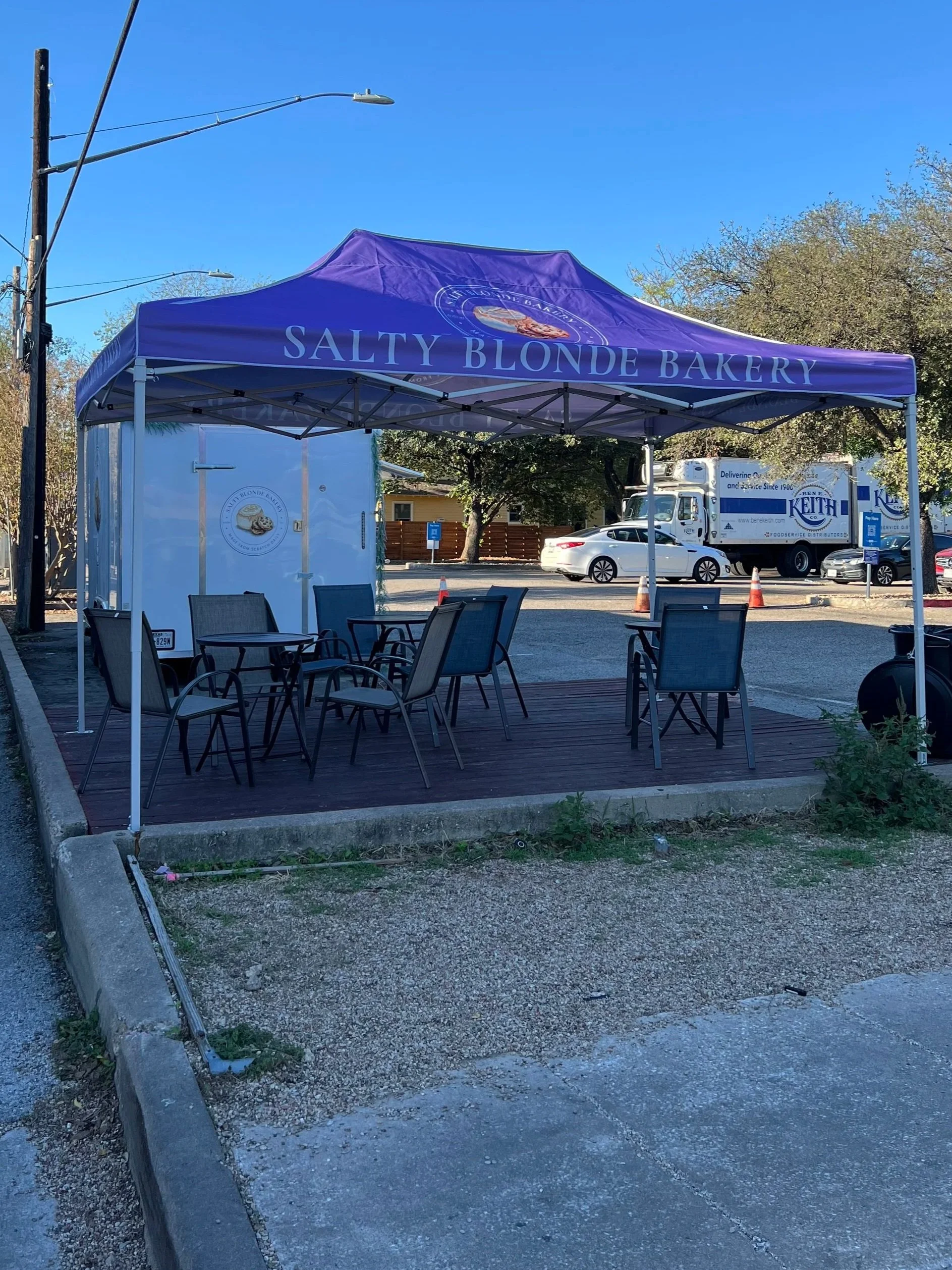 Outdoor seating area under a blue canopy with the words 'Salty Blonde Bakery,' with chairs and tables, in a parking lot with cars and trucks in the background.
