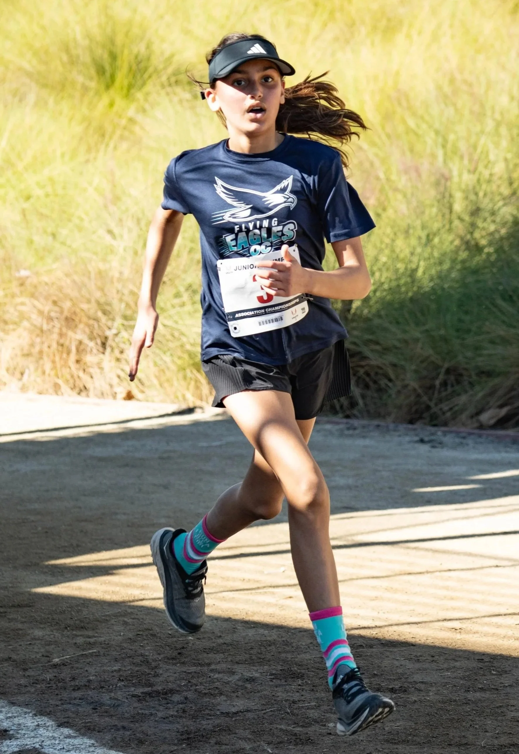 A young girl running outdoors on a dirt path, wearing a black visor, navy t-shirt with a graphic, black shorts, colorful striped socks, and running shoes, participating in a race.