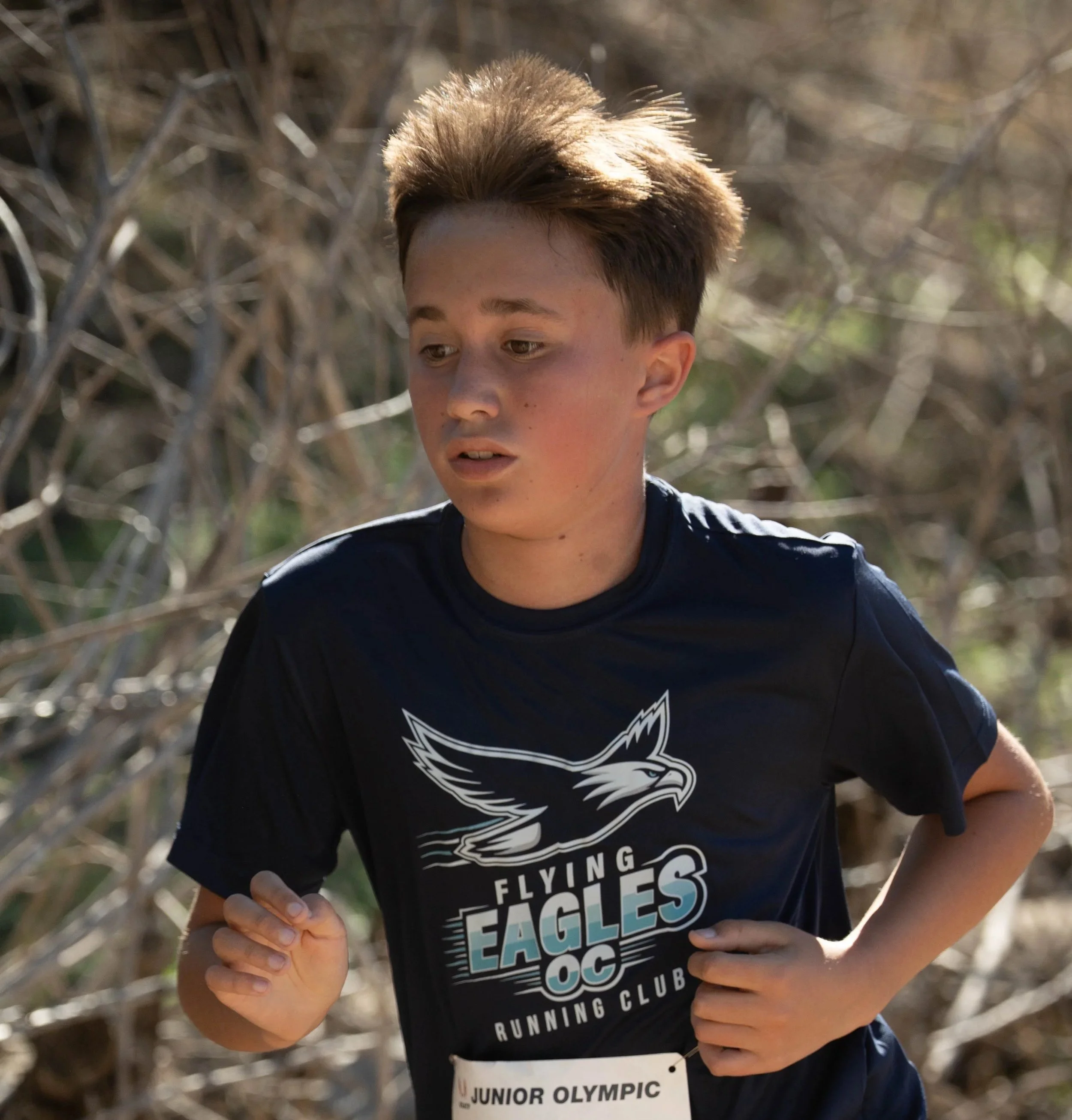 A young boy running outdoors wearing a black t-shirt with a flying eagle logo and the text 'Flying Eagles OC Running Club'.