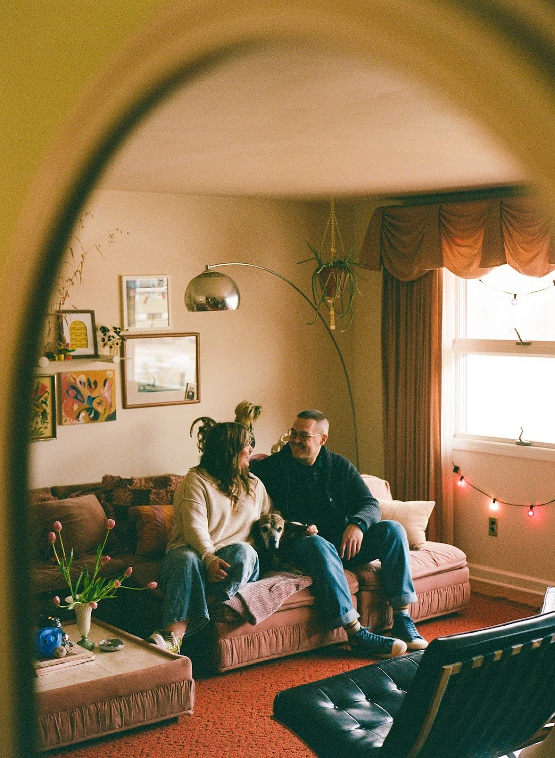 A couple sitting on a pink sofa with a small dog in a cozy living room. The woman is wearing a cream sweater, and the man is in a dark sweater. There are framed pictures and colorful artwork on the wall, a silver arc floor lamp, a hanging plant, and a window with curtains and string lights.