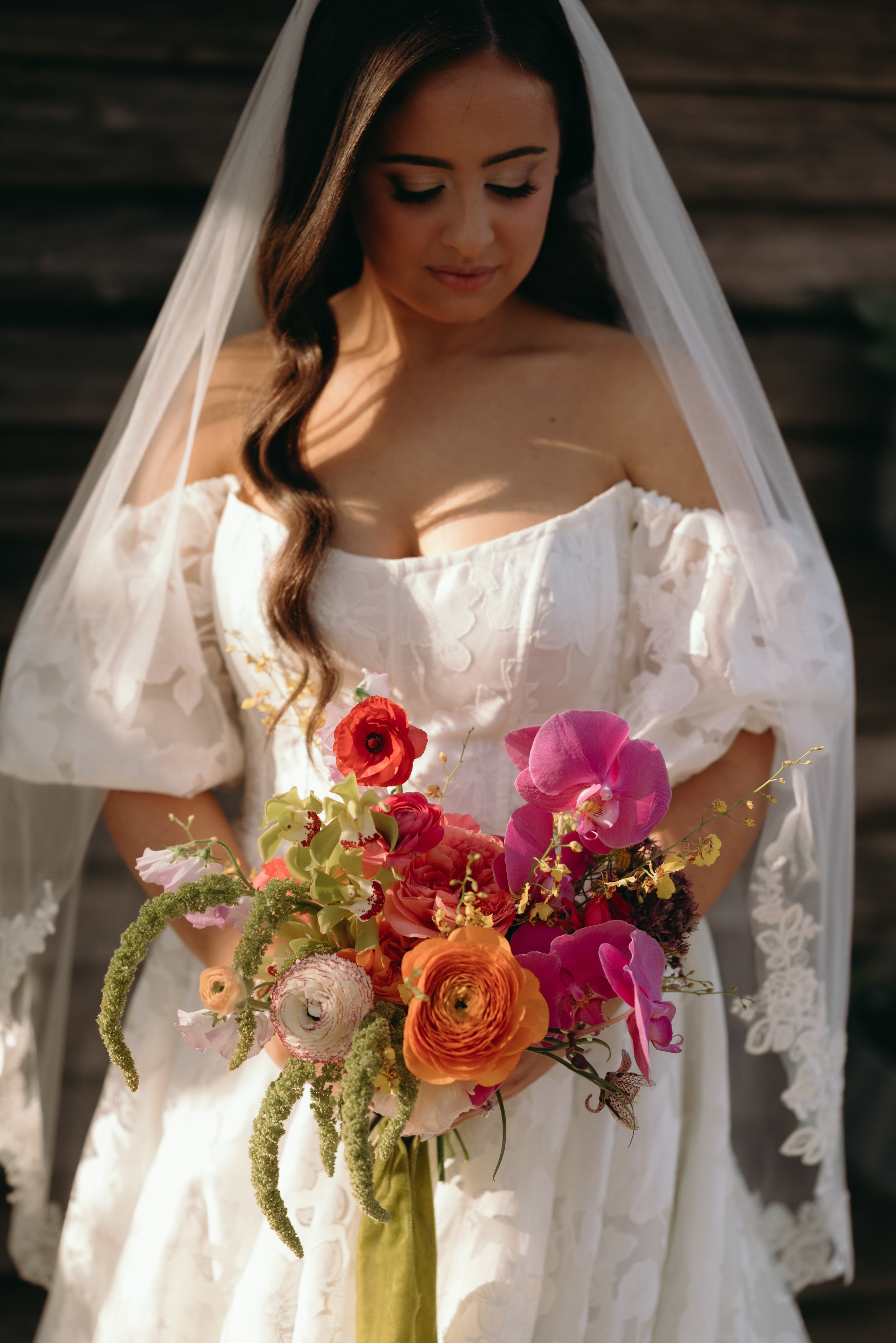A woman in a white wedding dress holding a colorful bouquet of flowers, with a veil over her hair, standing outdoors in front of a wooden background.
