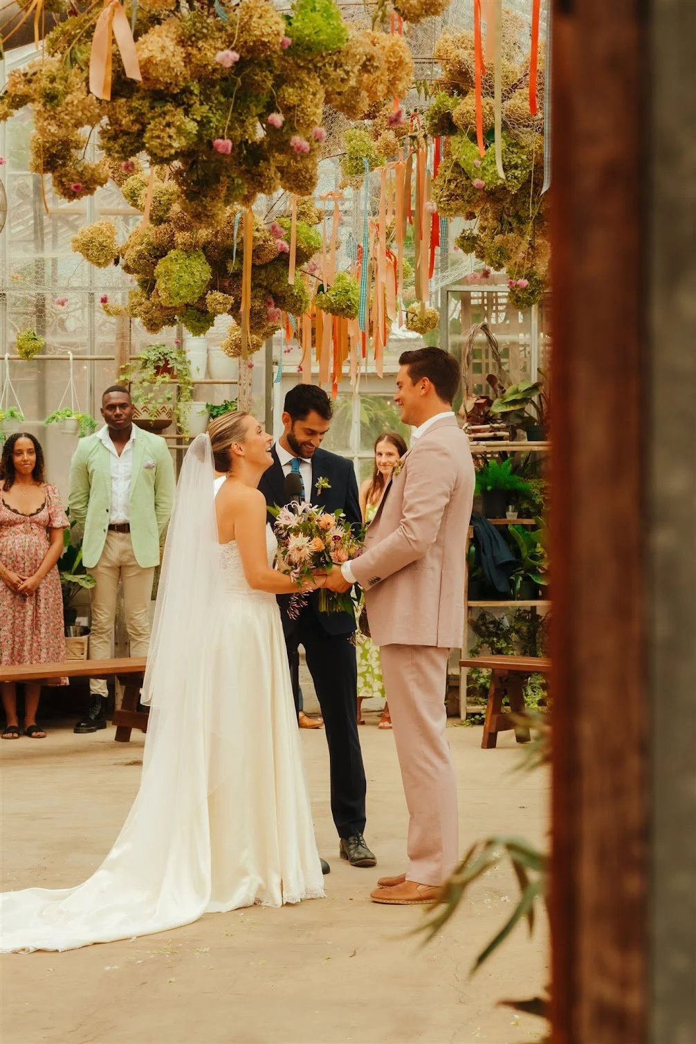 A wedding ceremony with a bride and groom exchanging vows in a greenhouse decorated with hanging flowers and ribbons, surrounded by friends and family.