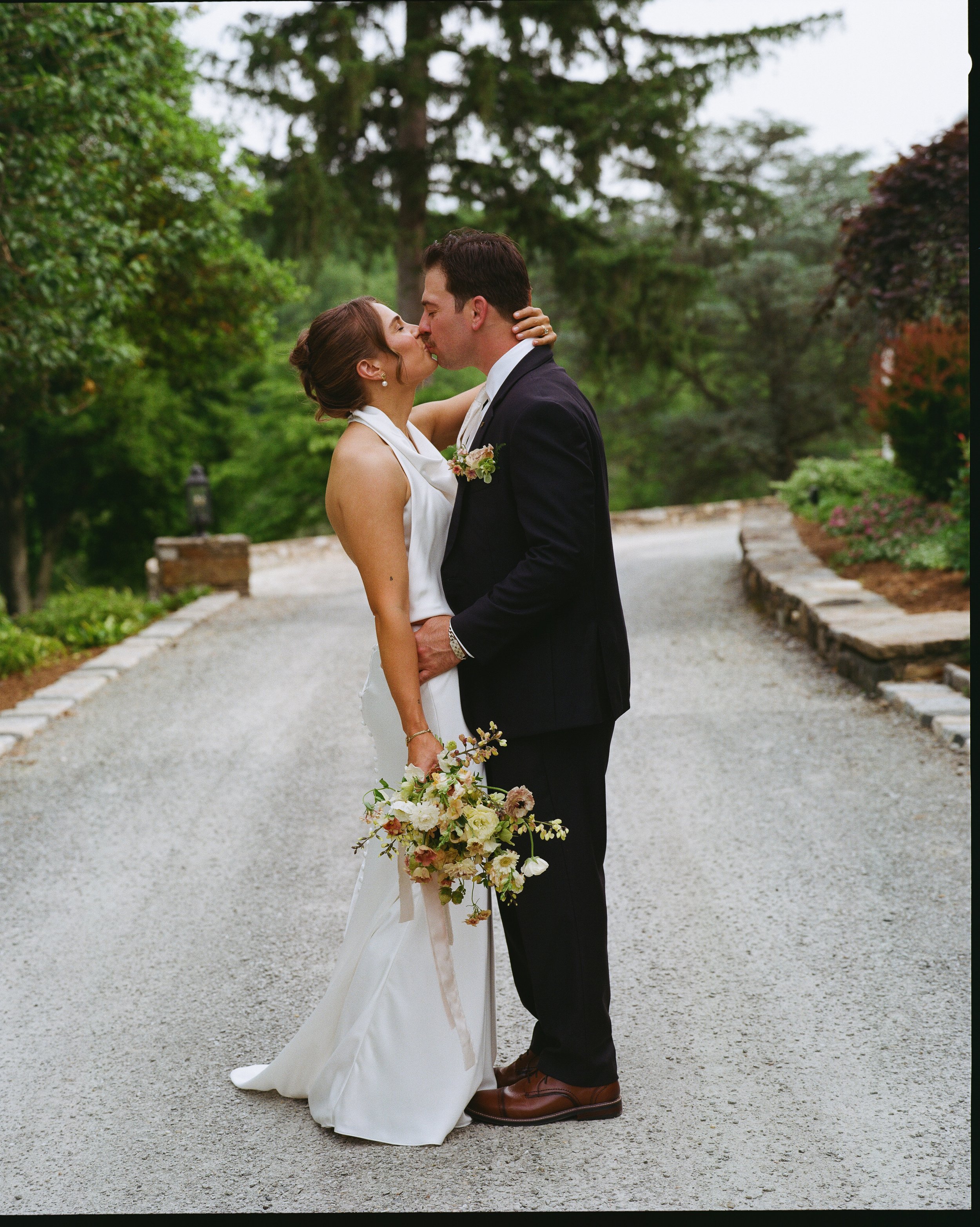 A bride and groom sharing a kiss outdoors. The bride is dressed in a white wedding gown, holding a bouquet of flowers, and the groom is wearing a dark suit with a boutonniere. They are standing on a gravel path with lush green trees in the background