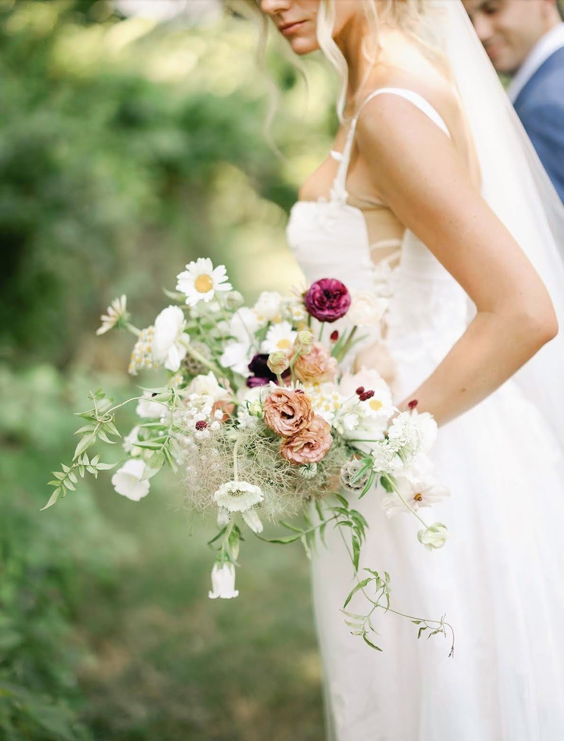 A bride in a white wedding dress holding a bouquet of assorted flowers, standing outdoors with greenery in the background.