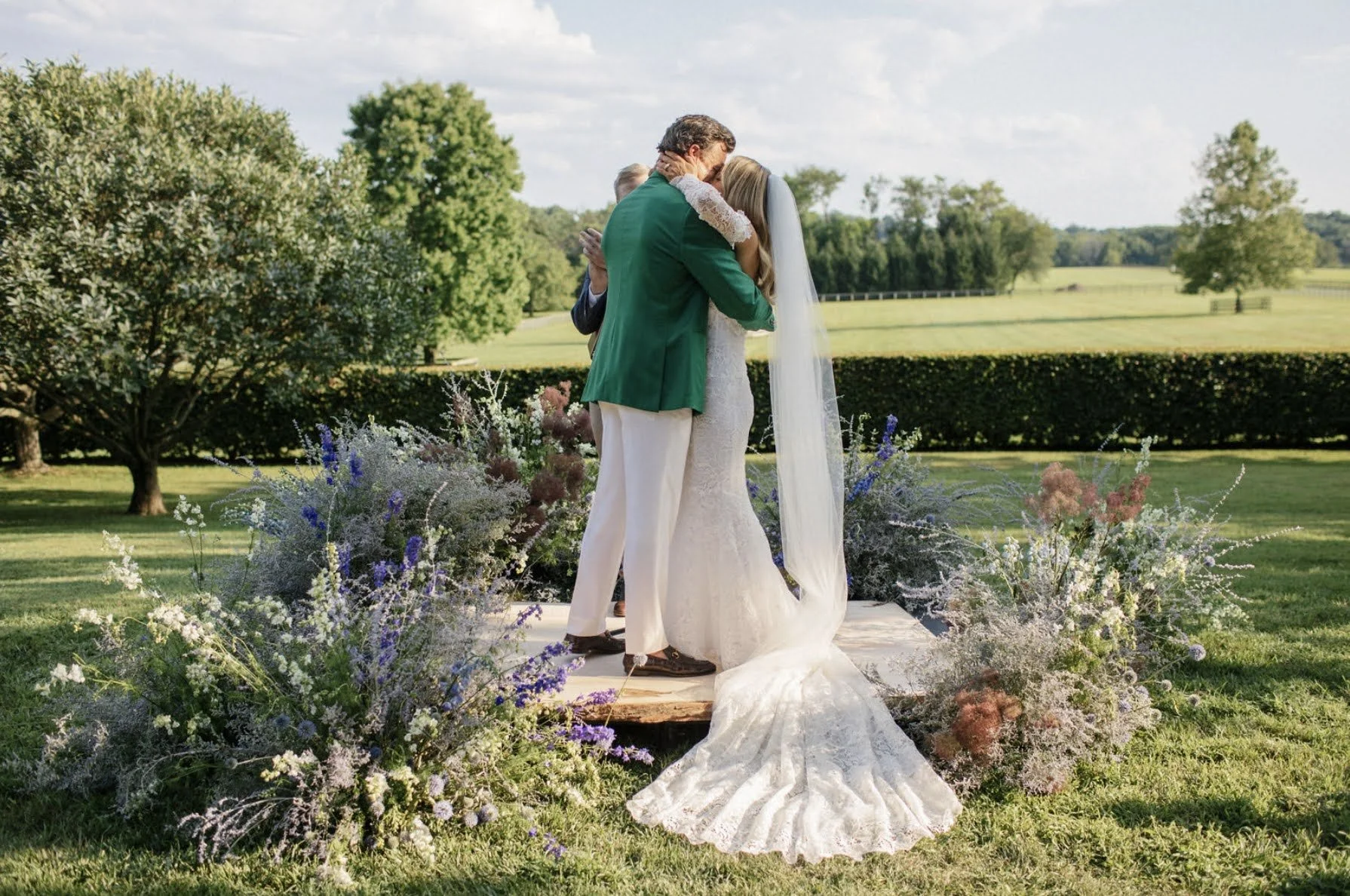 A couple dressed in wedding attire sharing an embrace during an outdoor wedding ceremony surrounded by floral arrangements, with a scenic green landscape and trees in the background.