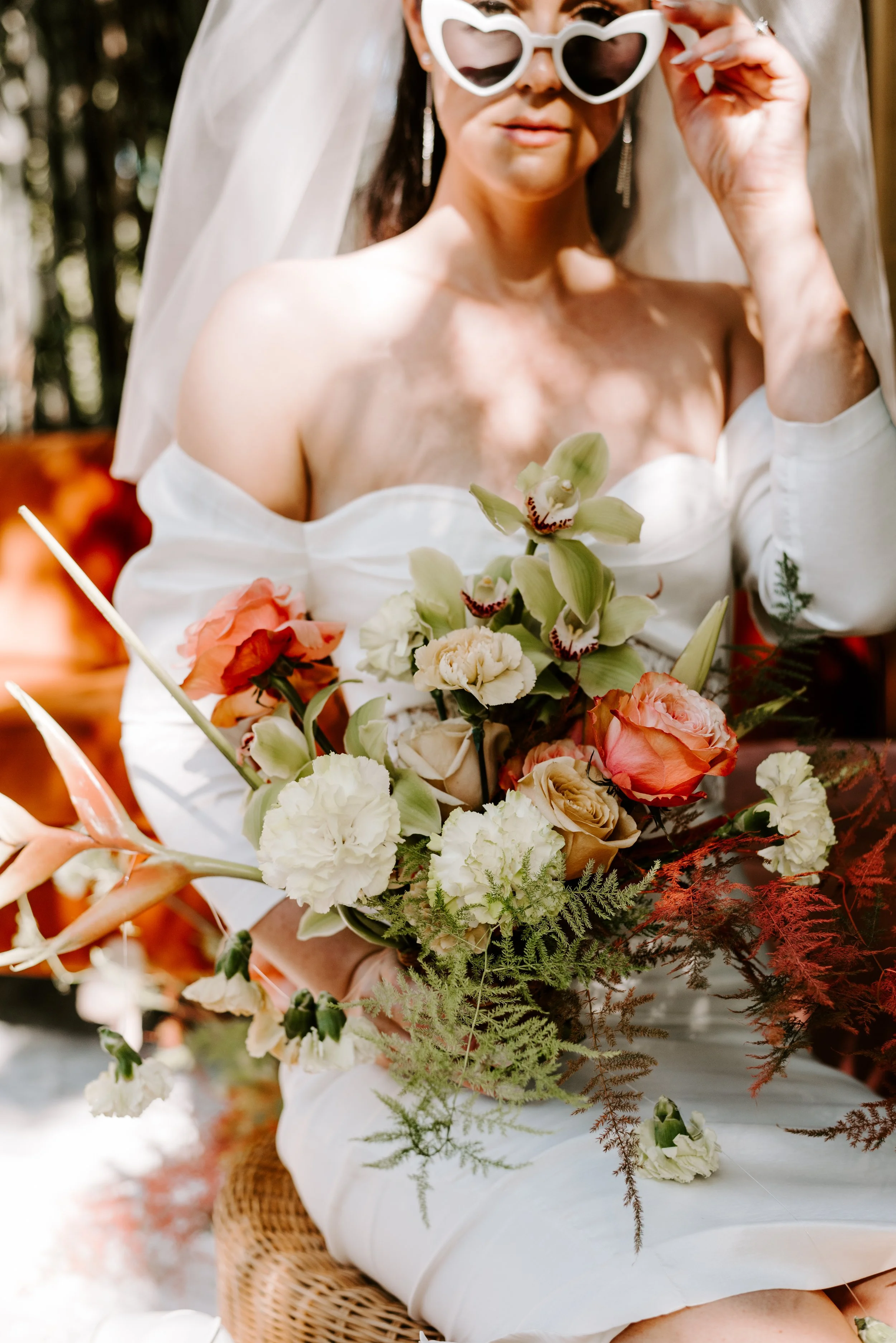A woman in a white off-shoulder dress holding a bouquet of flowers, wearing heart-shaped sunglasses and a veil, standing outdoors surrounded by flowers and plants.