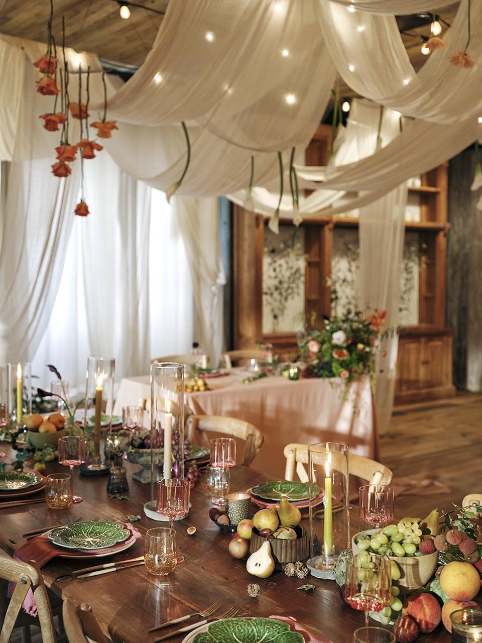 A decorated dining table with candles, bowls of fruit, and place settings in a rustic, cozy room with draped fabric and floral arrangements.