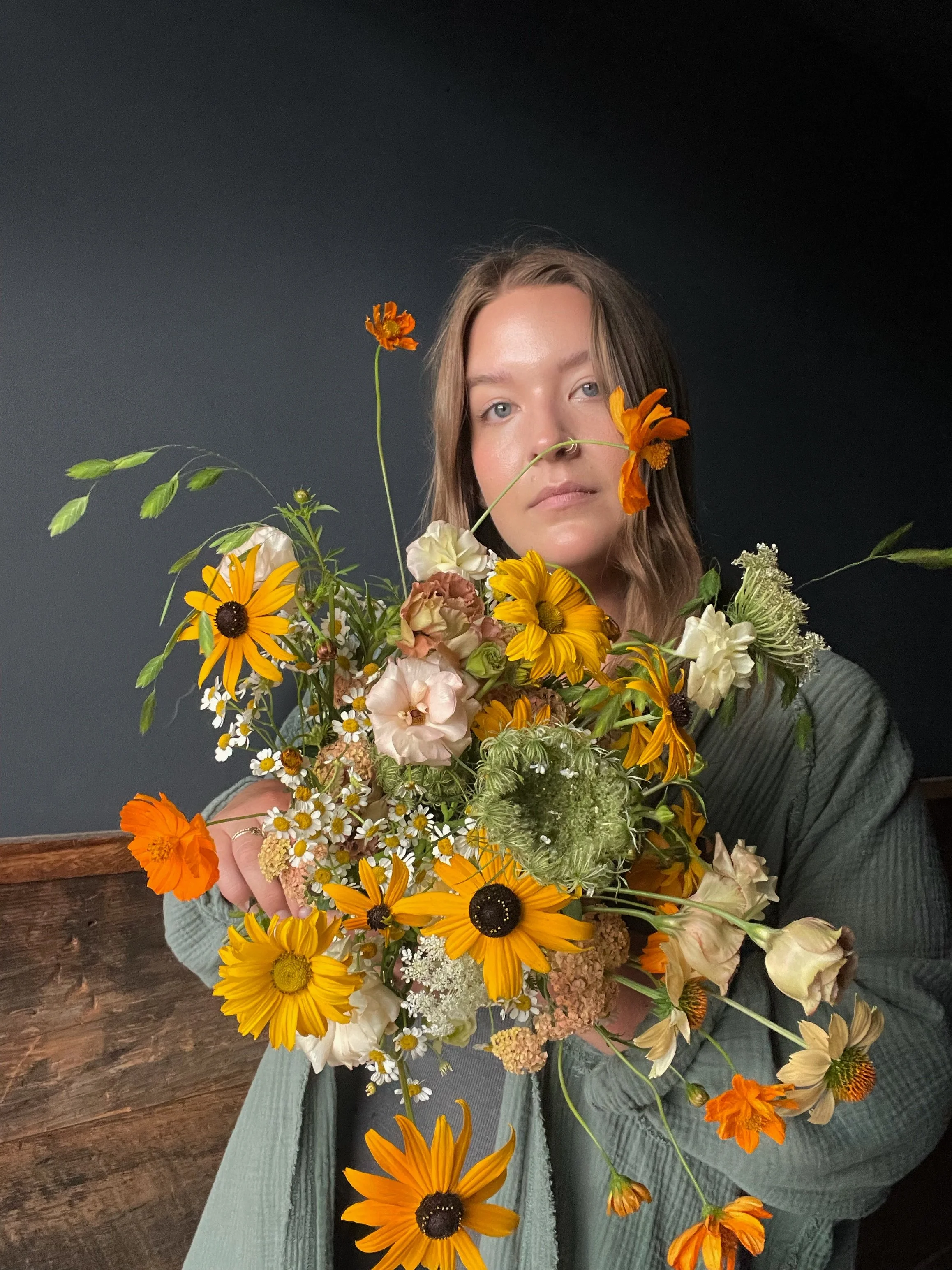 Person holding a large bouquet of various colorful flowers including orange, yellow, and cream blooms, with a plain dark background.