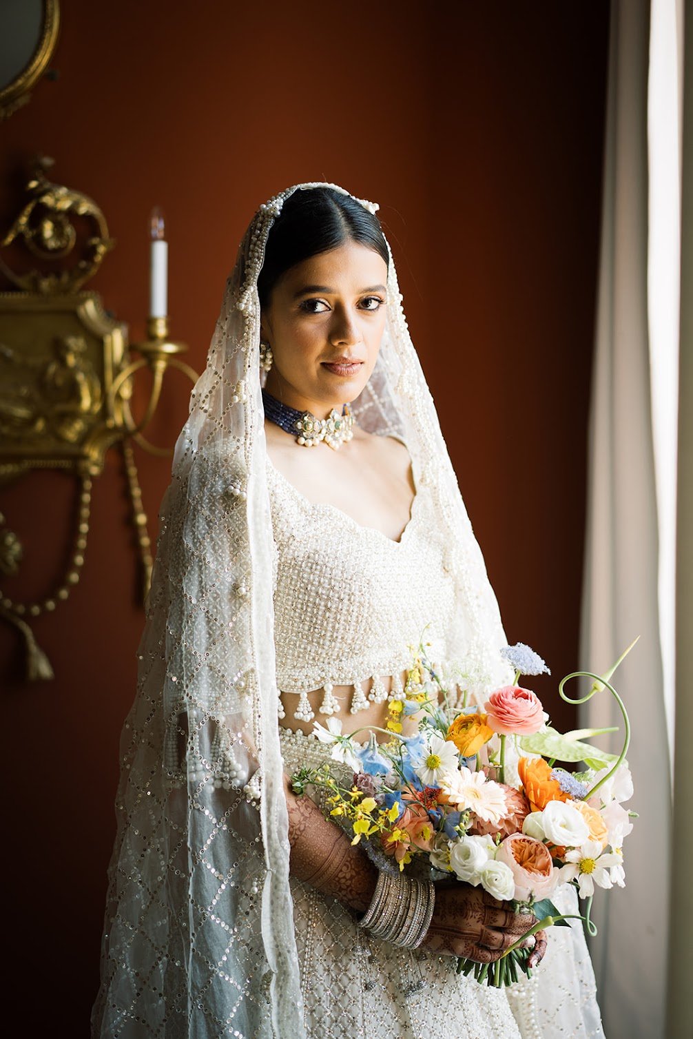 A bride standing indoors near a window, dressed in traditional wedding attire with intricate jewelry, holding a colorful bouquet of flowers, with a red wall and a gold wall sconce in the background.