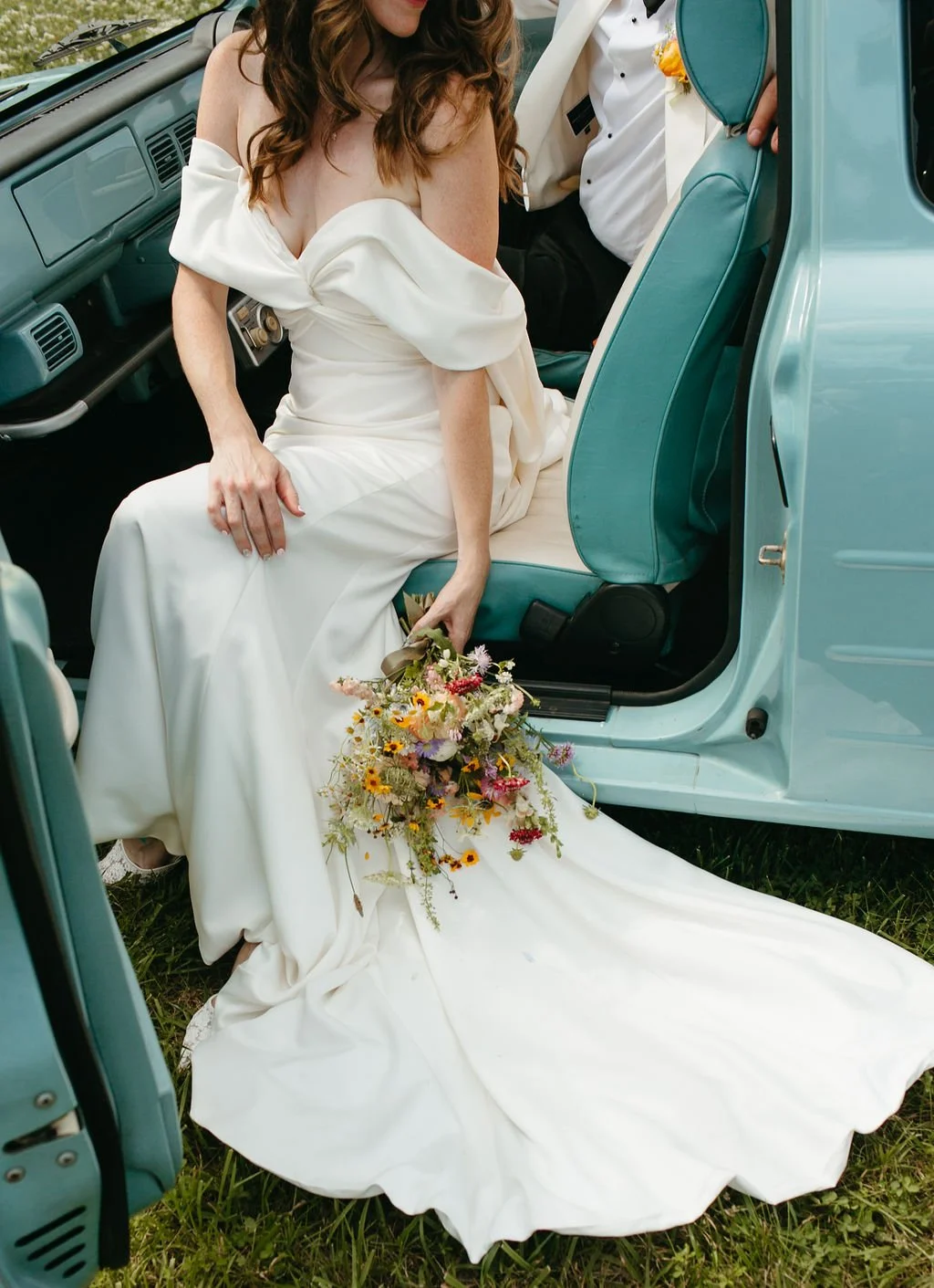 A bride in a white wedding dress sitting on the edge of an open vintage car door, holding a bouquet of colorful flowers, with her dress trailing on the grass.