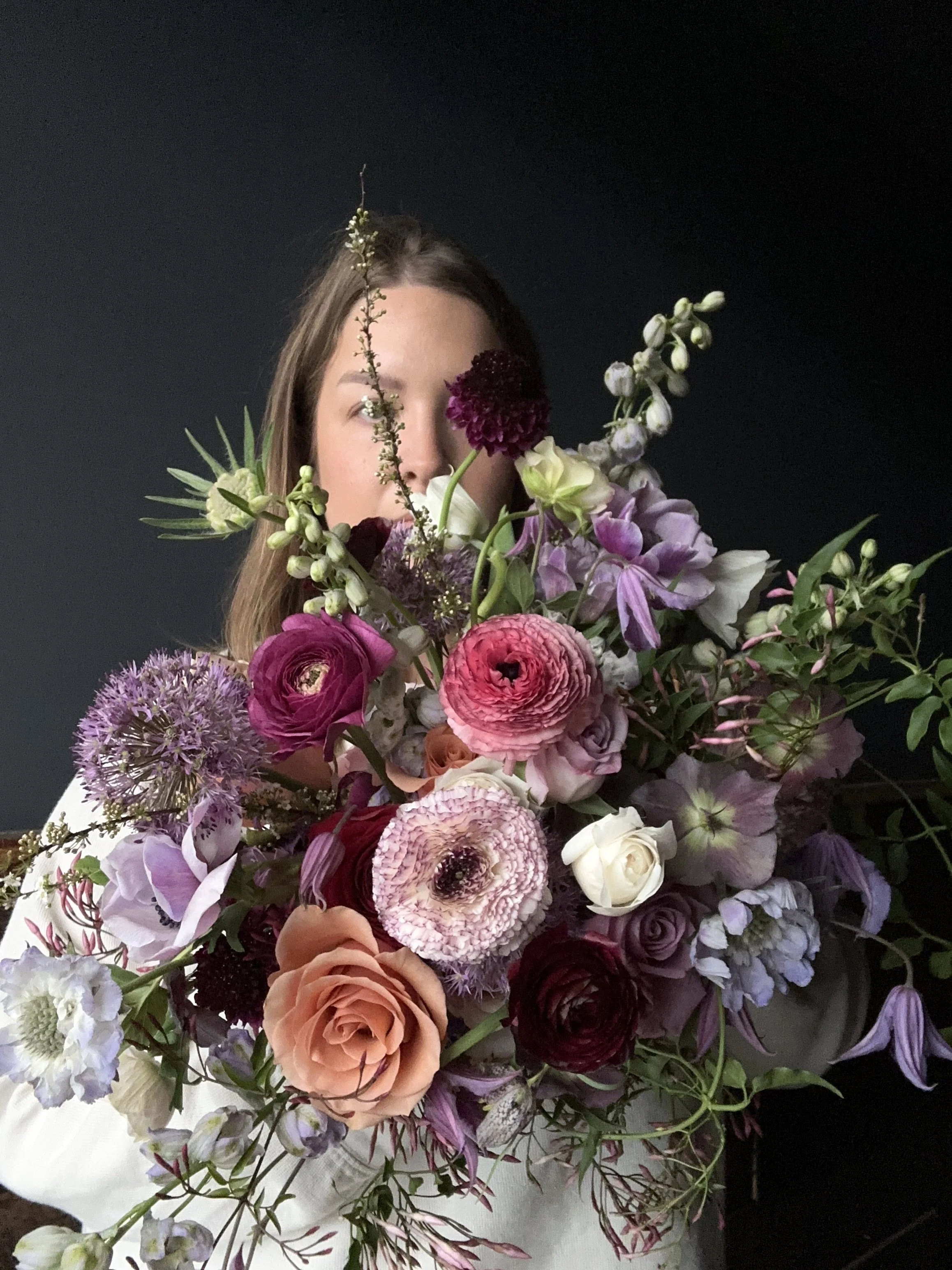 A woman holding a large bouquet of various colorful flowers, including roses, peonies, and other blooms, partially obscuring her face against a dark background.