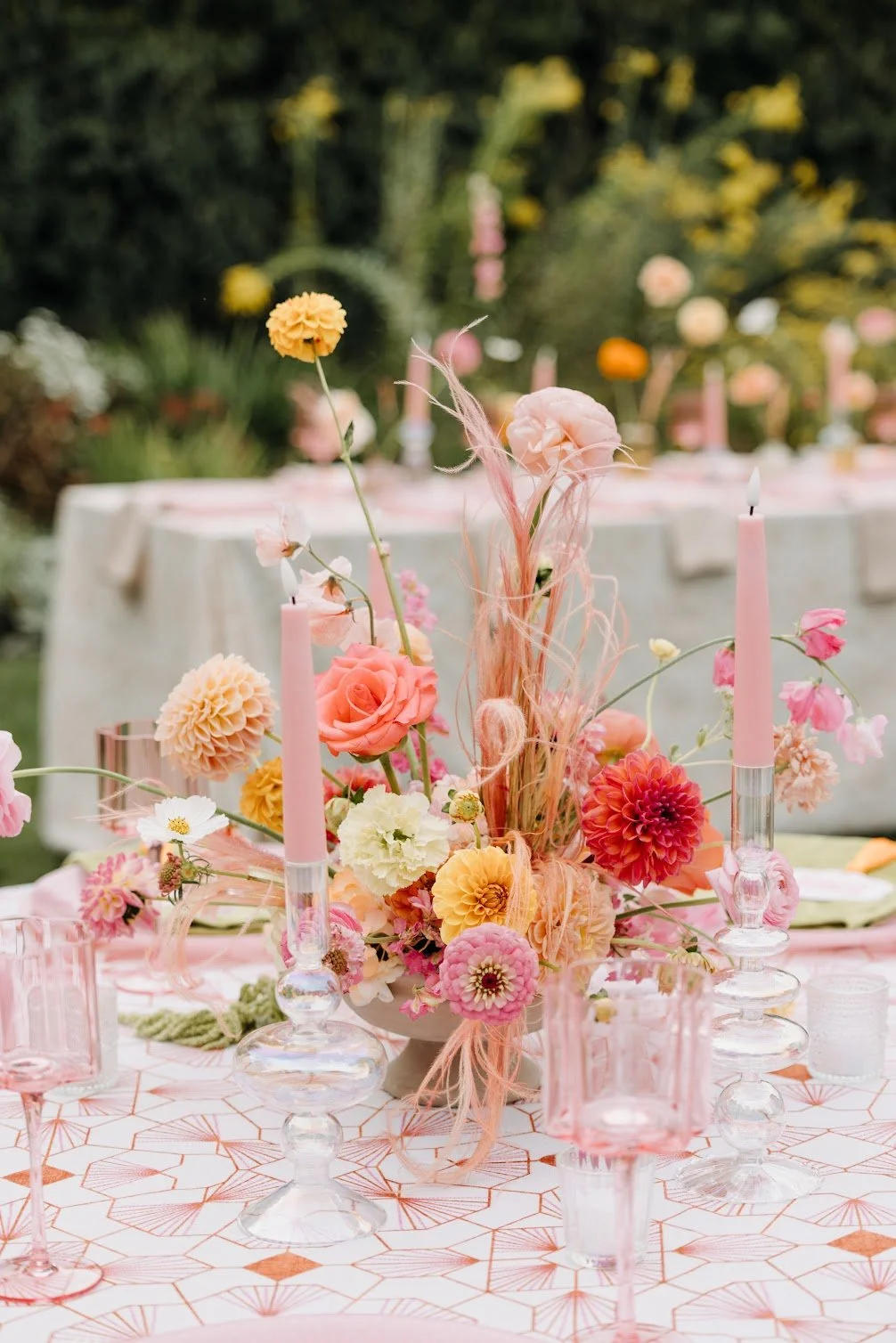 A table decorated with a floral centerpiece consisting of pink, peach, and yellow flowers, pink candles, and pink glassware, set outdoors with a blurred background of trees and greenery.