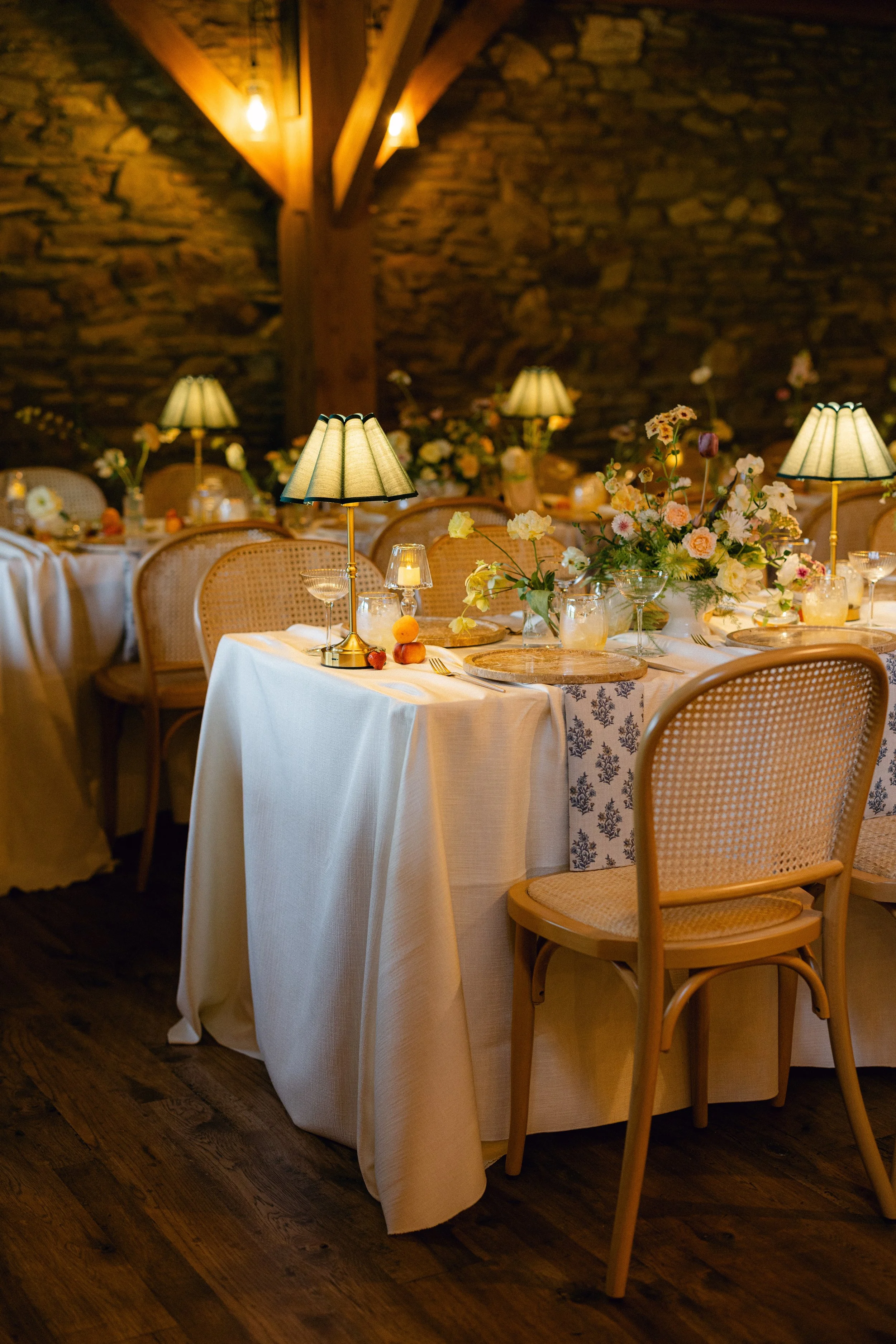 Elegant dining room with tables set with white tablecloths, floral centerpieces, and wicker chairs. Small lamps with green shades provide warm lighting, and a rustic stone wall with wooden beams is in the background.