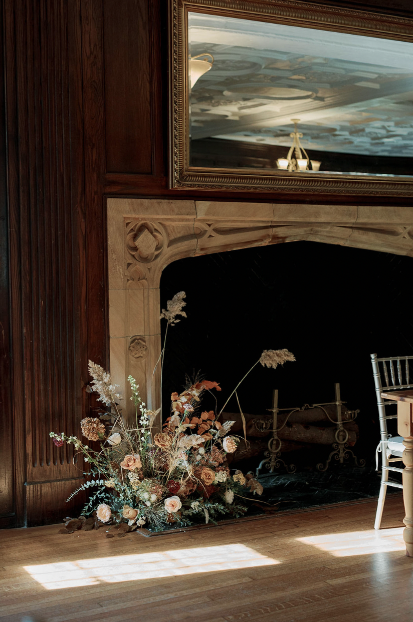 A floral arrangement with pink and white flowers and dried grasses placed in front of a fireplace in a wood-paneled room with a large mirror and a chandelier reflected in it.