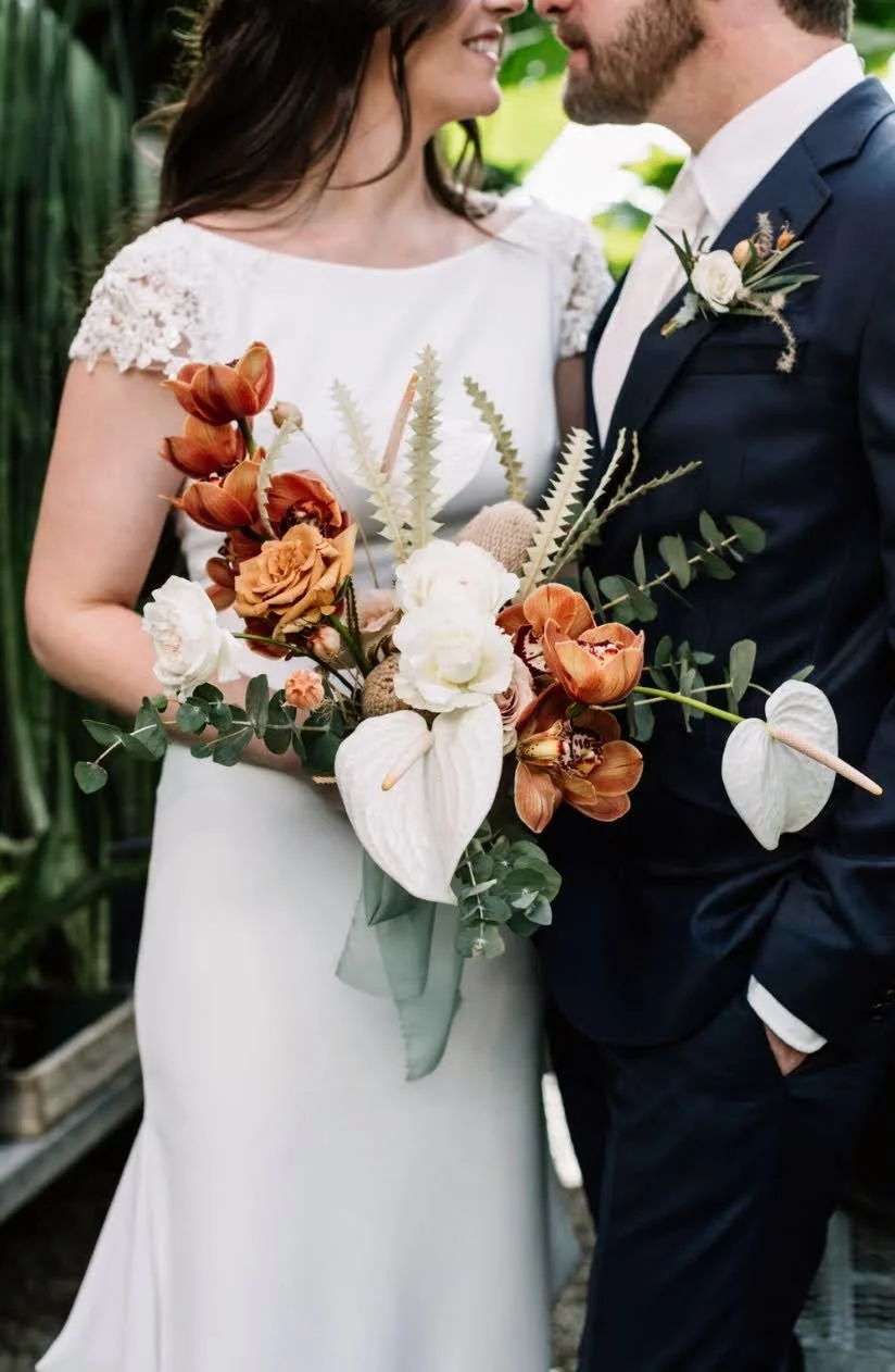 A bride and groom on their wedding day, smiling and facing each other. The bride is holding a large bouquet with white, orange, and brown flowers, as well as greenery. The groom is wearing a dark suit with a boutonniere. They are outdoors with lush g