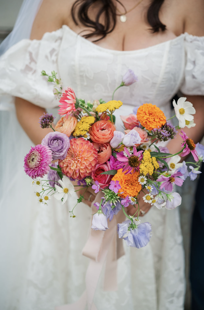 A bride holding a colorful bouquet of various flowers including pink, purple, white, yellow, and orange blooms, while wearing a white wedding dress with lace details.