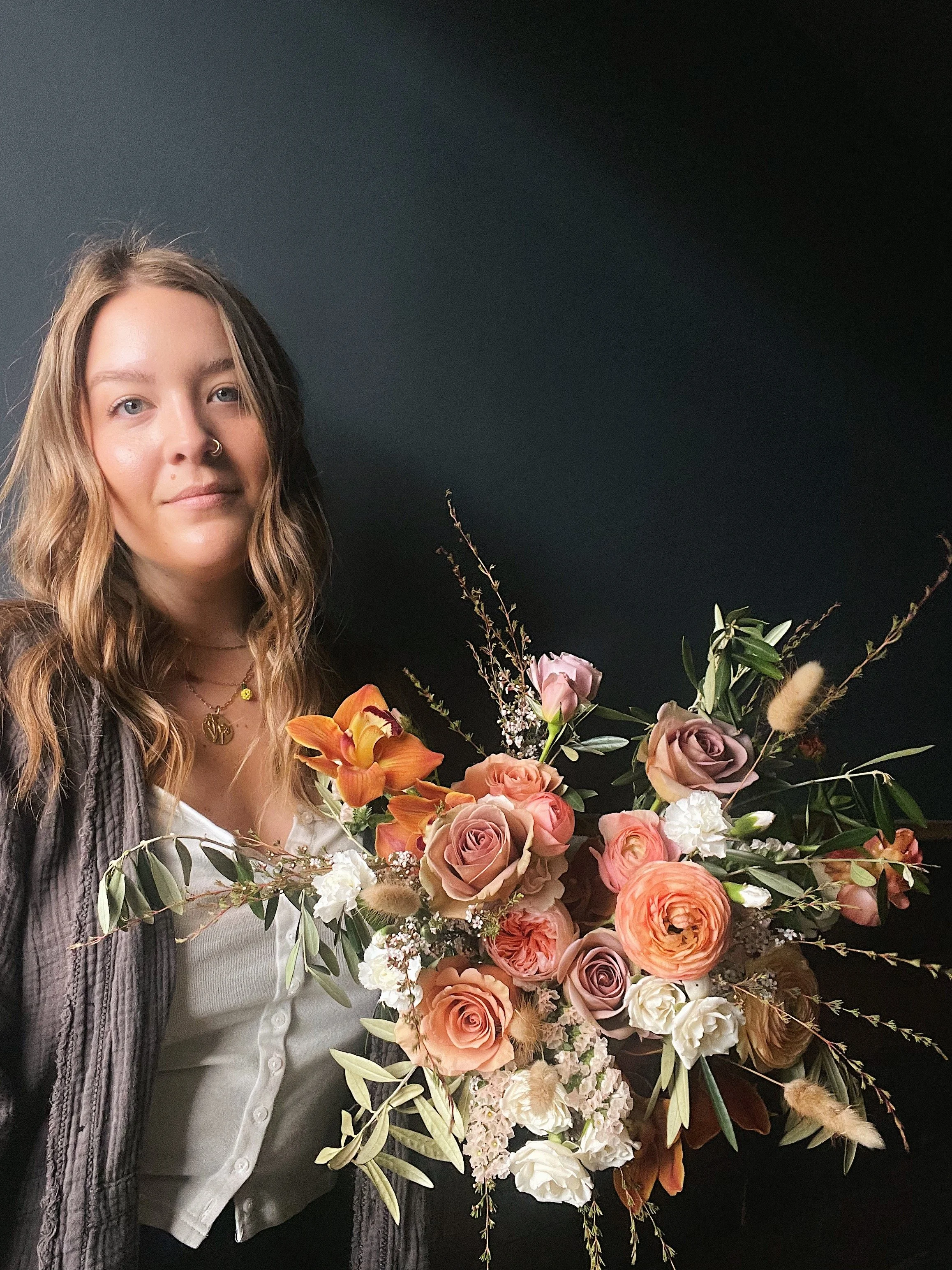 A woman with wavy light brown hair and a nose ring, wearing a white shirt and layered necklaces, holding a large bouquet of mixed roses, peonies, and greenery against a dark background.