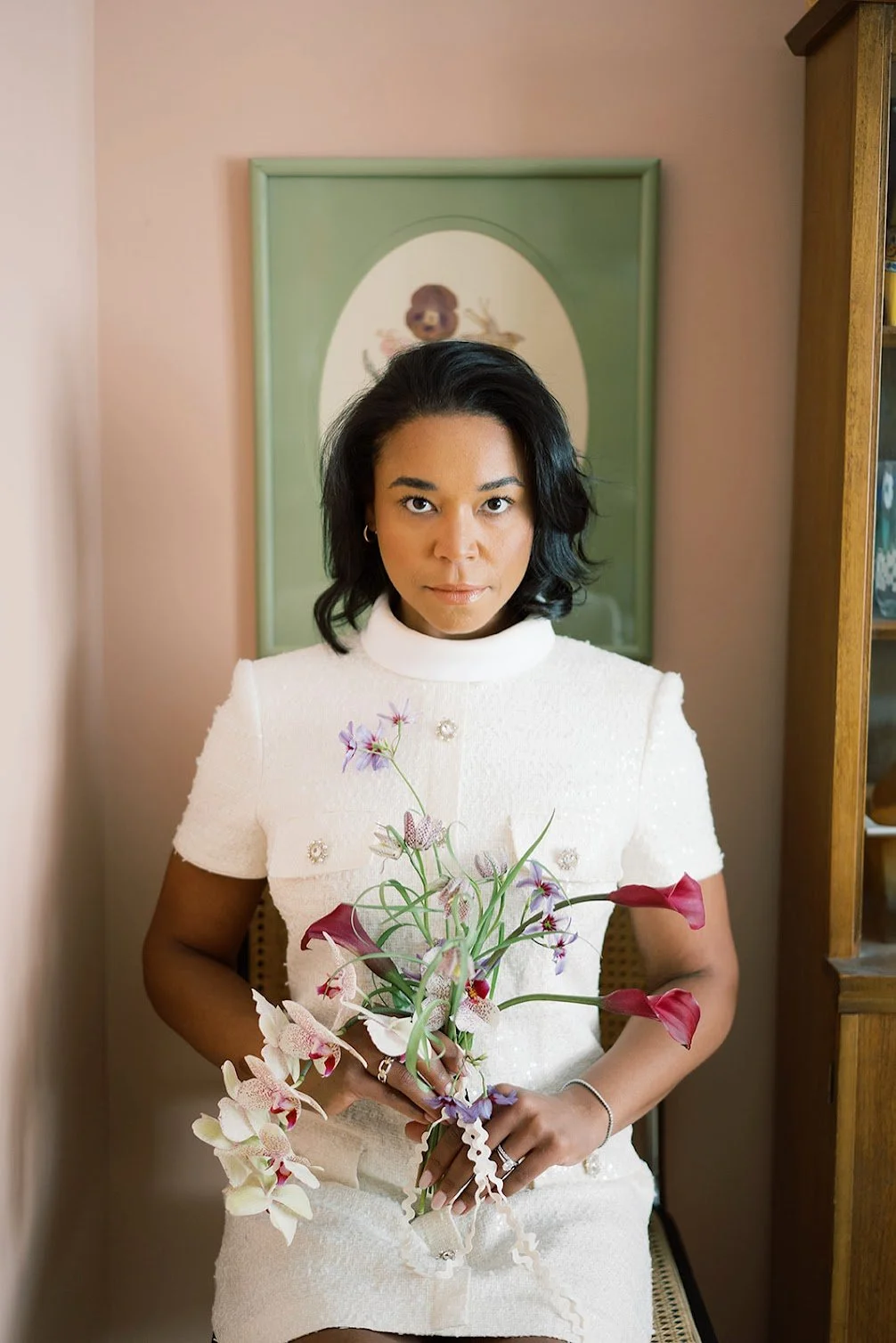 Woman with black hair holding a flower arrangement with orchids and calla lilies in a room with a pink wall and a framed picture of a bear in the background.
