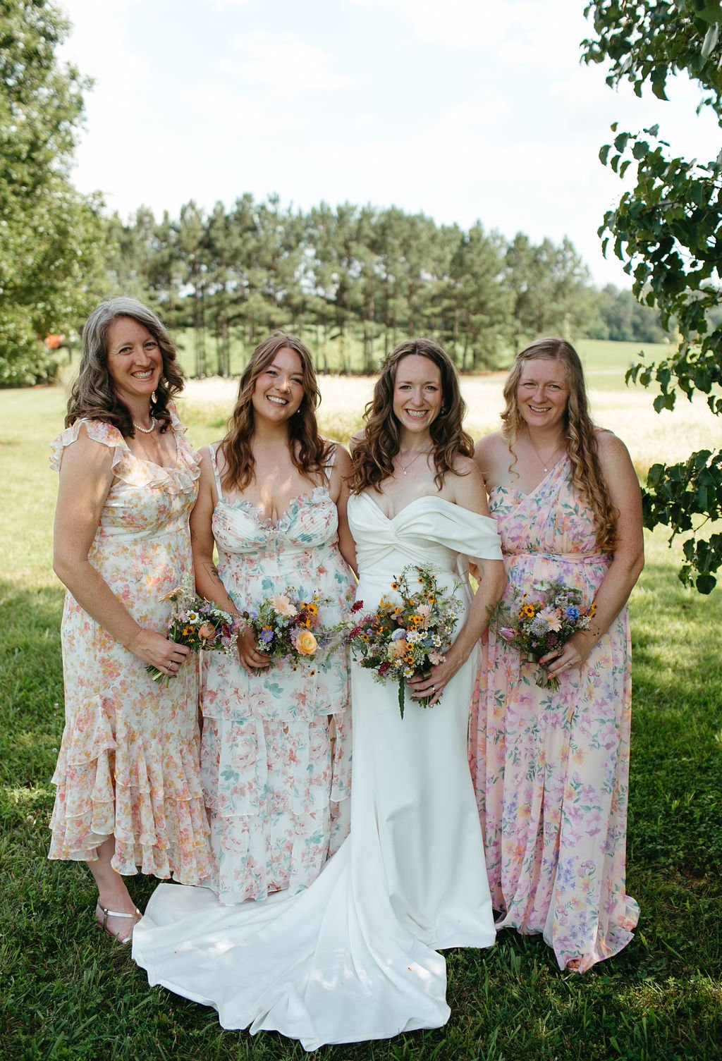 Four women standing outdoors on a grassy field, dressed in floral dresses, holding bouquets of flowers, smiling, with trees and a clear sky in the background.