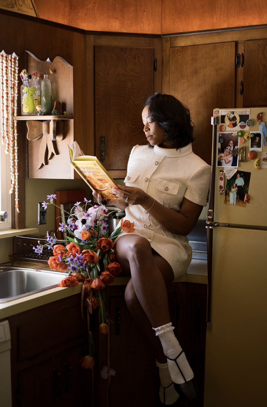 A woman sitting on a kitchen counter reading a cookbook, surrounded by a bouquet of orange and purple flowers, with a wooden kitchen interior and a refrigerator decorated with magnets and photos.