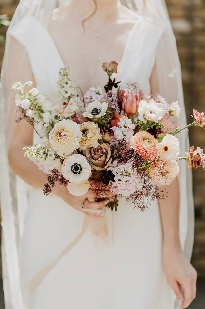 A bride holding a bouquet of pastel-colored flowers including roses, anemones, and ranunculus, in front of a blurred background.