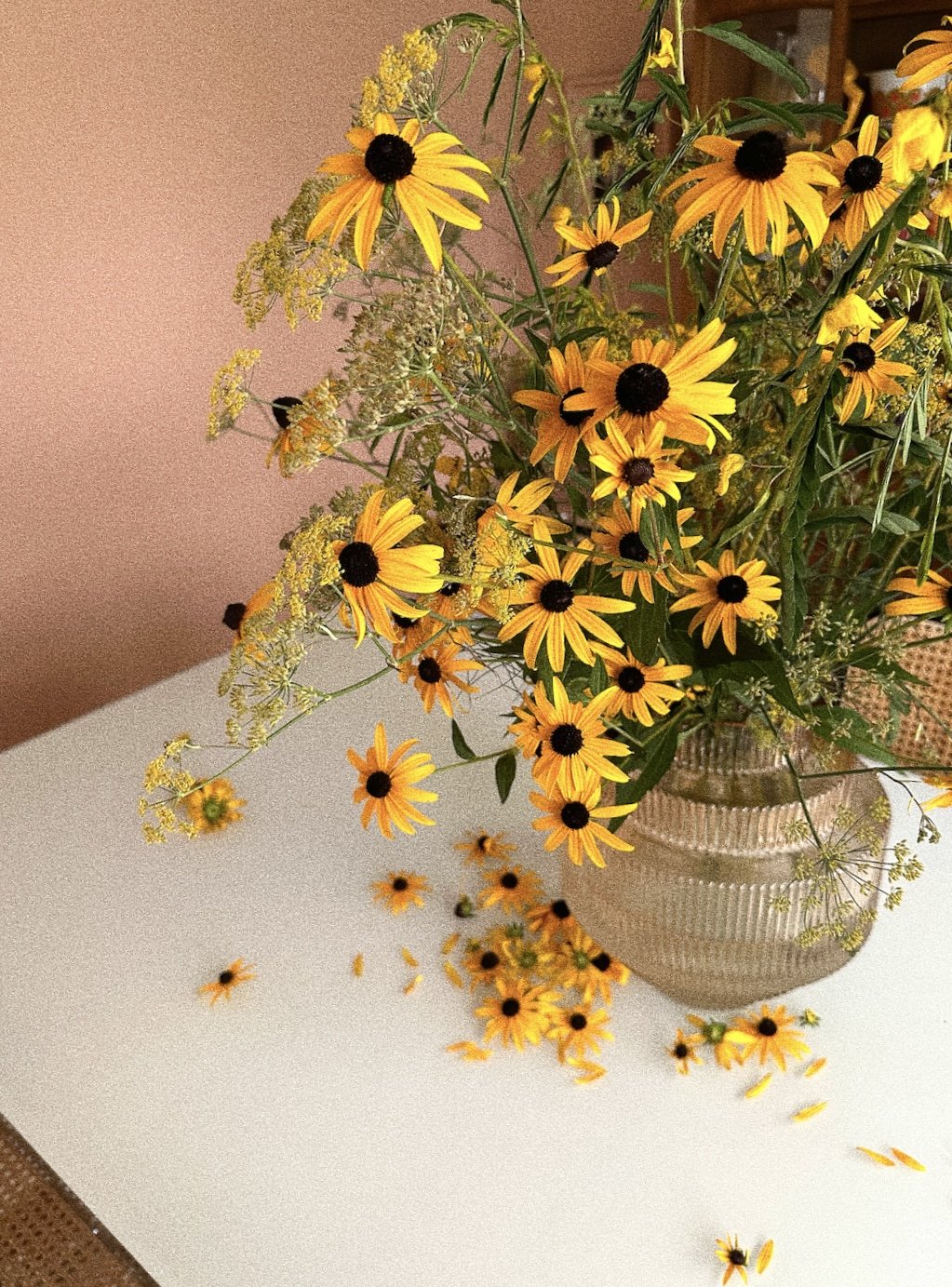 A bouquet of yellow flowers with dark centers in a glass vase on a white surface, some flowers spilling onto the table.