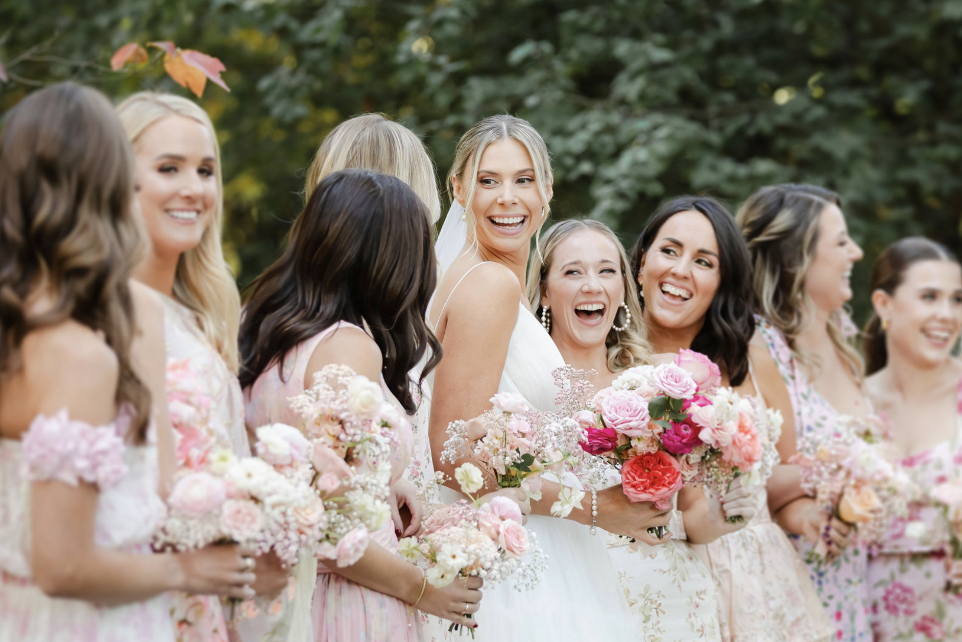 Group of women in dresses holding bouquets of pink and white flowers, standing outdoors with green trees in the background, smiling and laughing during a wedding celebration.